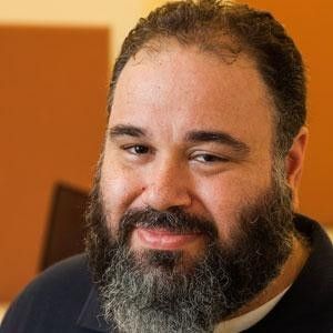 A man with a magnificent, salt-and-pepper beard and a warm, subtle smile gazes directly at the viewer. His dark hair is slightly receding, and his eyes convey a friendly, approachable demeanor, perhaps hinting at a hidden joke. He wears a dark polo shirt. The background is a simple, warm brown wall, suggesting an indoor, possibly office or classroom setting, which provides a neutral backdrop to his prominent facial hair. The sheer volume and distinguished grey streaks in his beard give him a wise, almost wizard-like appearance, making one wonder if he's about to share ancient secrets or just a really good dad joke. His expression suggests he's ready for either.
