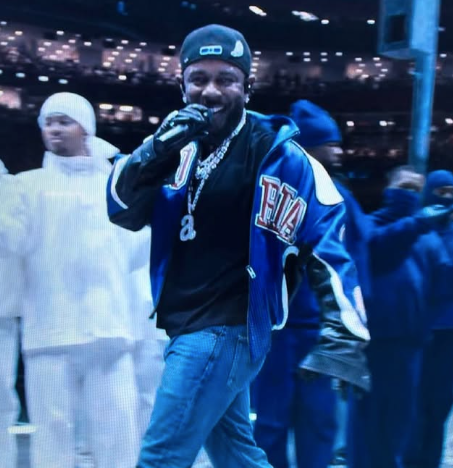 The image captures an energetic performer, likely from a live music event, dressed in a stylish ensemble featuring a vibrant jacket and accessories that sparkle under the stage lights. His demeanor is lively and engaging, possibly interacting with an enthusiastic audience, which adds an electric atmosphere to the scene.
In the background, a crowd is visible, with some individuals dressed in white, perhaps indicating a theme or concept for the performance. The lighting is dim, with twinkling lights creating a sense of excitement and anticipation. This setting suggests a concert or major event where the performer is the focal point, drawing the audience's attention. Overall, the image radiates a fun and vibrant vibe, typical of live performances, where the connection between the performer and the audience is palpable.