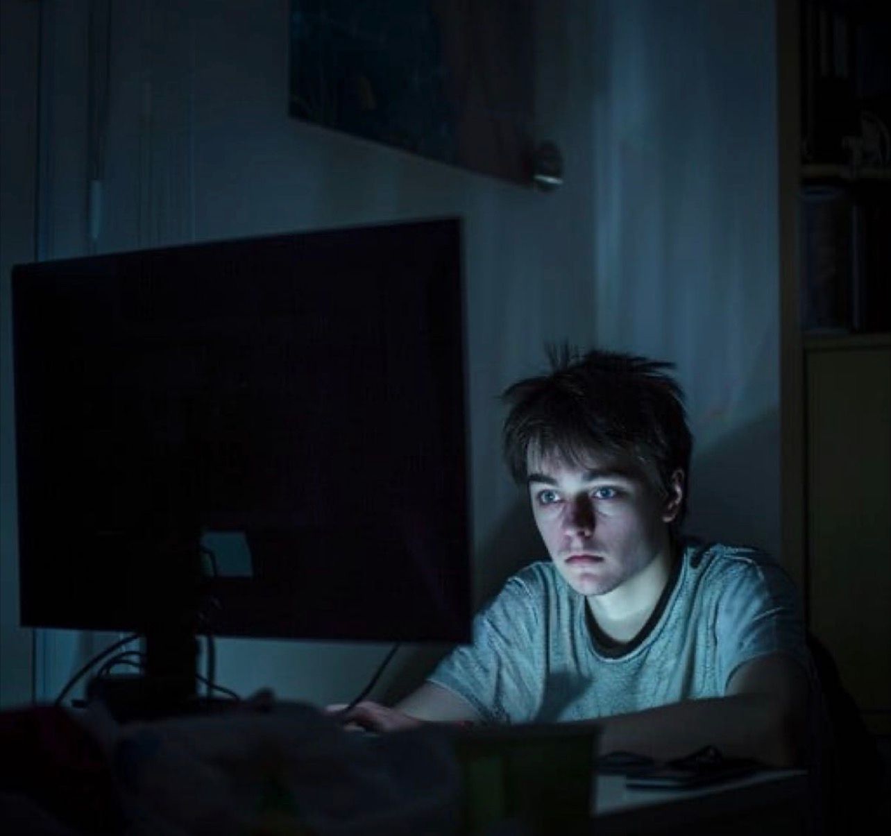 The image shows a young person with short, dark hair sitting in front of a computer monitor in a dimly lit room. The person is wearing a light gray t-shirt and appears to be focused on the screen. The light from the monitor illuminates their face, casting shadows on the surrounding area. The background is mostly dark, with some indistinct objects and furniture visible.