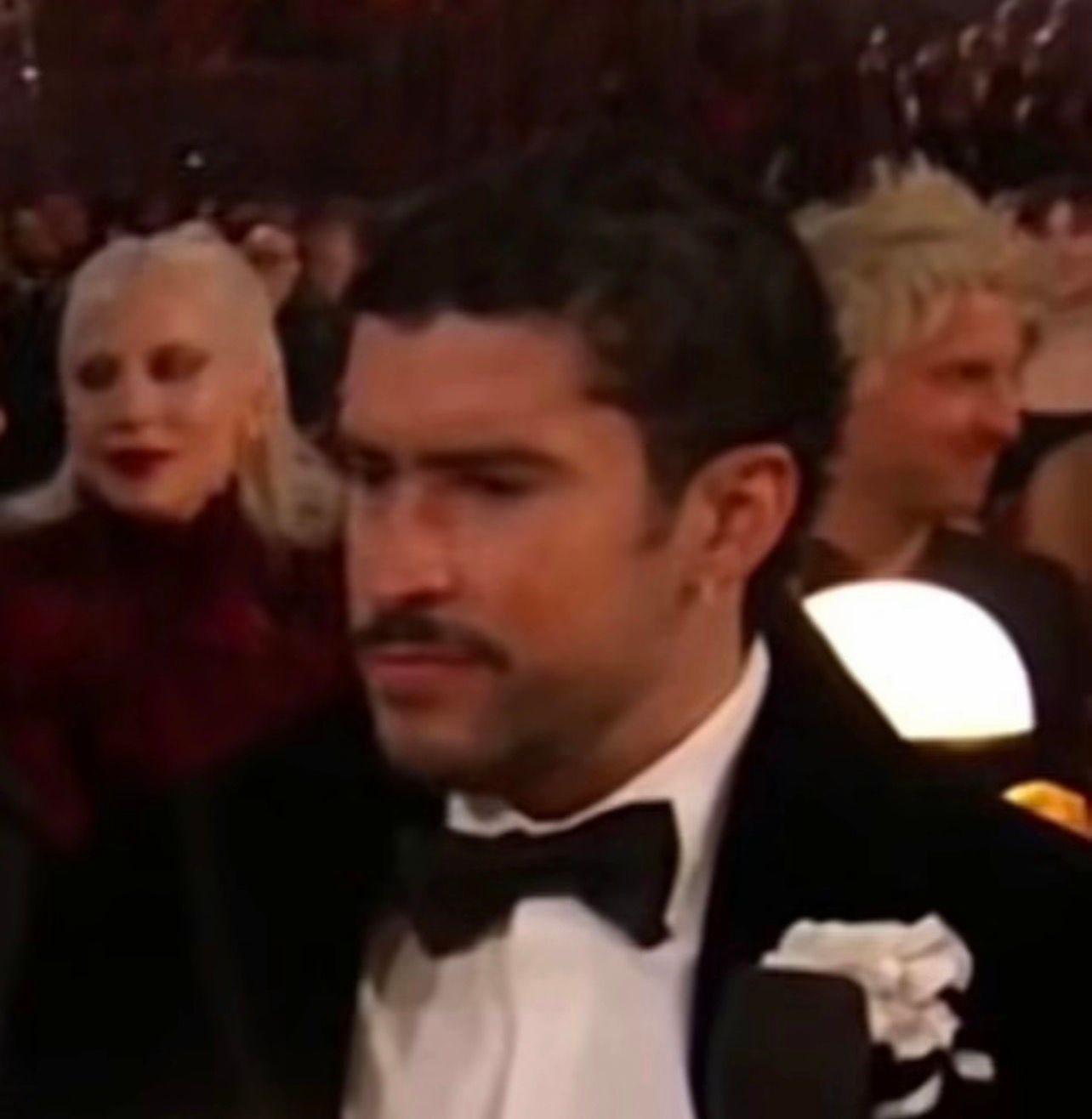 The image features a man in formal attire, wearing a tuxedo with a bow tie, exuding a serious demeanor. His facial expression suggests deep contemplation or perhaps a moment of distraction, as he gazes slightly to the side. He has a neatly groomed mustache, adding to his distinguished appearance. In the background, a woman with platinum blonde hair and a striking red lip is visible, seemingly engaged in conversation, which adds a layer of social interaction to the scene. The setting appears to be a glamorous event, likely an awards ceremony, given the elegant attire and the ambiance. The soft lighting, along with the blurred figures behind the man, enhances the focus on him while hinting at the celebratory atmosphere around. The contrast between his serious expression and the lively background creates a humorous juxtaposition, as if he’s pondering the meaning of life while surrounded by glitz and glamour.