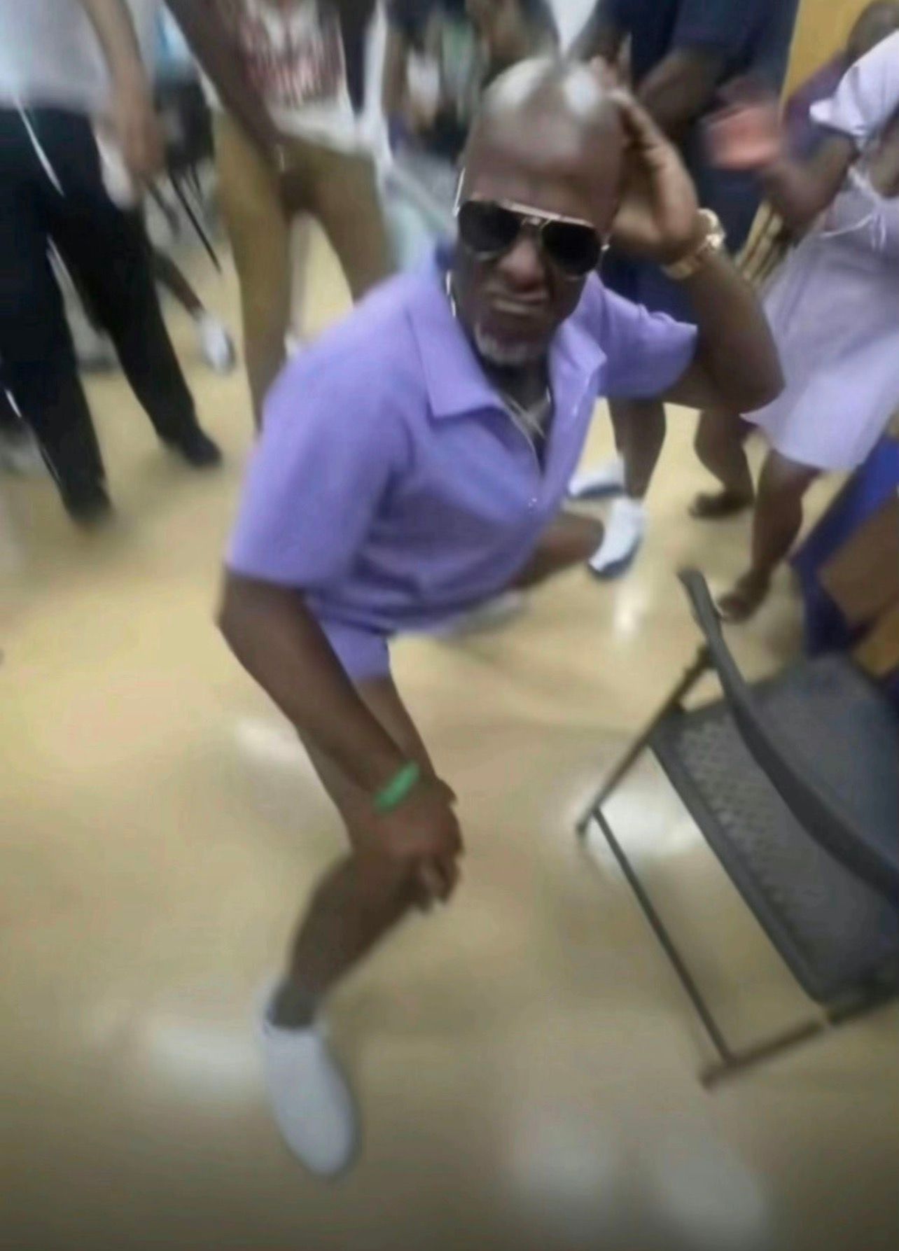 The image features a man in a vibrant purple outfit, complete with sunglasses and a playful pose that suggests he’s in the midst of dancing or having fun. His demeanor is confident and carefree, displaying a sense of enjoyment. The background is filled with people, though they are somewhat blurred, indicating a lively atmosphere, likely at a social gathering or celebration.
The setting, characterized by a simple indoor space with a light-colored floor, adds to the casual and festive vibe of the scene. The overall context suggests a joyful event, perhaps a party or reunion, where everyone is engaged in fun activities.
Humorously, the man appears to be striking a pose as if he’s the star of the show, adding a playful touch to the gathering. His outfit and confident demeanor seem to say, “I’m here to party, and everyone else should take notes!”