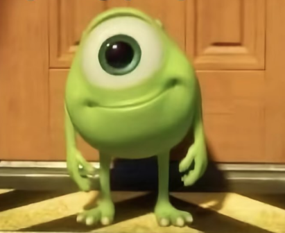 The image shows a green, one-eyed animated character standing in front of a wooden door. The character has a large single eye centered on its round head, a small smile, and two arms and legs with three fingers and toes on each hand and foot. The character is standing on a floor with a patterned design. The lighting suggests it is indoors.