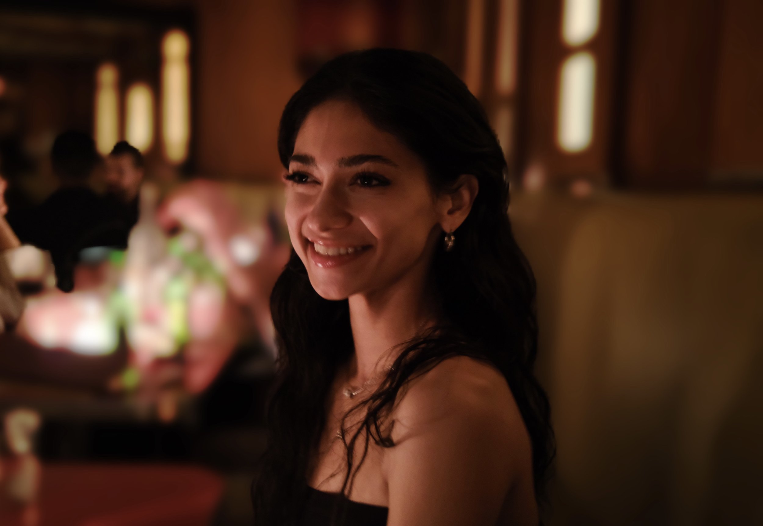 The image features a woman with long, dark hair, smiling warmly as she engages with her surroundings. Her demeanor is joyful and inviting, suggesting a moment of connection or happiness. The soft, ambient lighting creates a cozy atmosphere, enhancing her smile and highlighting her features.
In the background, blurred figures and hints of colorful lights suggest a lively social setting, possibly a restaurant or a gathering, evoking feelings of camaraderie and celebration. The overall ambiance hints at a relaxed evening with friends or loved ones, where laughter and good conversation are likely taking place.
The blurred background complements the foreground, emphasizing the subject’s expression and drawing attention to her moment of joy. The scene captures the essence of shared experiences, making it relatable and warm-hearted.