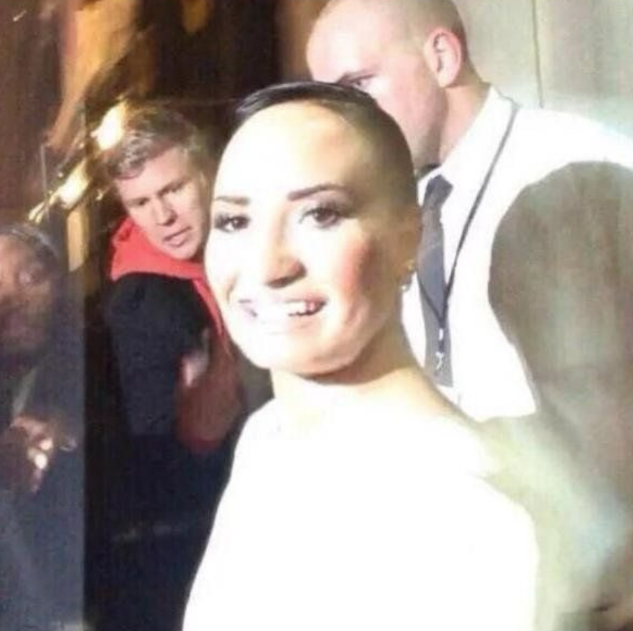 The image presents a manipulated photograph featuring a person strongly resembling Demi Lovato in the foreground, smiling broadly with a cheerful and confident demeanor. Her most striking feature is a significantly exaggerated, smooth forehead, giving her a comically large, bald-appearing "fivehead." This distortion is the central humorous element. In the background, a man in a red hoodie with blonde hair peers over her shoulder, his face expressing clear surprise or bewilderment, seemingly reacting to the unusual sight. Further back, a bald man in a white shirt and dark tie, likely security, stands stoically. The background figures' reactions, especially the man in red, amplify the meme-like humor of the foreground subject's digitally altered appearance, creating a comical scene of exaggerated features and bewildered onlookers.