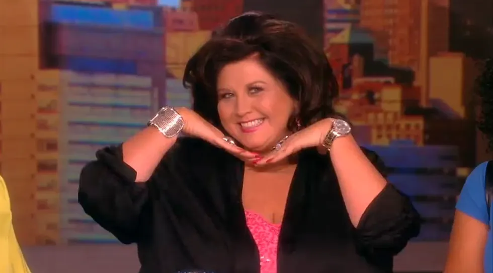 Abby Lee Miller, known for her role in Dance Moms, is captured in a television appearance, striking a surprisingly playful pose. She smiles broadly, her hands clasped under her chin, fingers adorned with rings and red nail polish, while sporting a large silver cuff and a gold watch. Her dark, voluminous hair frames a cheerful, almost mischievous expression, a humorous contrast to her typically stern demeanor on the show. She wears a black top over a sparkly pink sequined garment. The blurred, colorful background, resembling a stylized cityscape, is characteristic of a TV studio set, reinforcing that this is likely an interview or promotional segment. Her 'cute' pose, so different from her usual tough persona, adds a touch of unexpected humor to the image.
