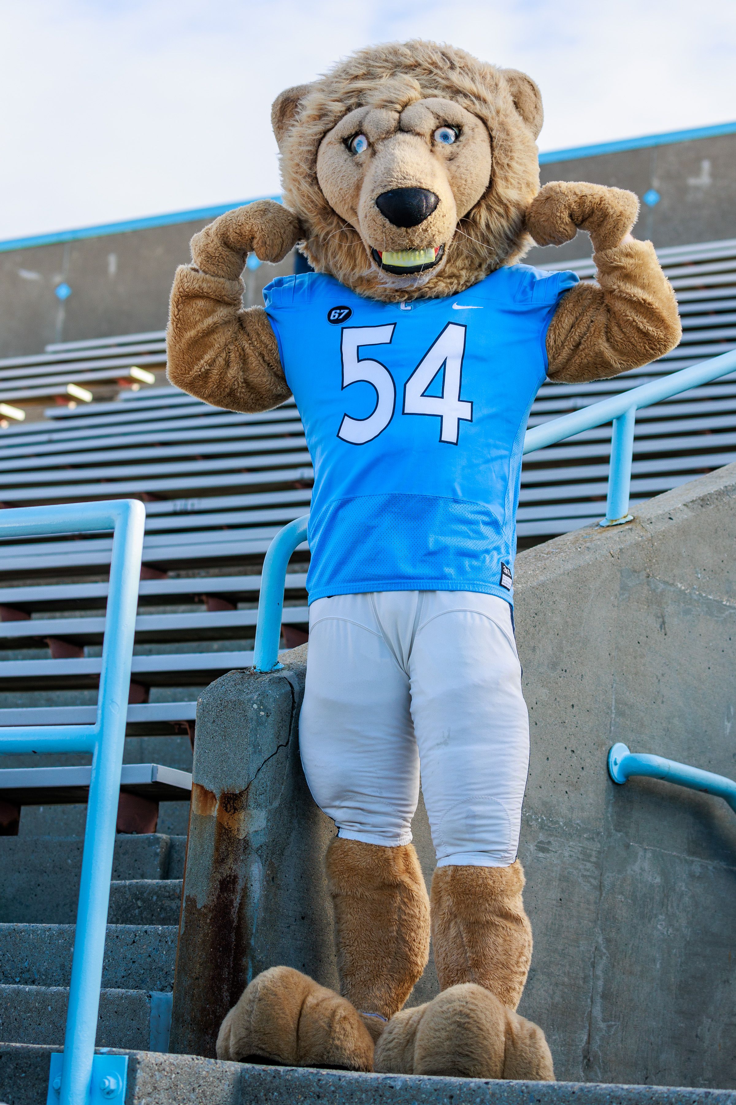 The image features a lion mascot, exuding an energetic and proud demeanor as it flexes its muscles, showcasing enthusiasm and team spirit. Clad in a blue jersey marked with the number "54," the mascot stands confidently on stadium steps, embodying the vibrant atmosphere of a sports event. In the backdrop, empty bleachers hint at the anticipation of fans, reinforcing a sense of community and excitement. The lion's exaggerated facial expression, complete with a wide grin and striking blue eyes, adds to the humor, making it a lively character ready to engage with the crowd. This scene encapsulates the essence of school pride and camaraderie, with the mascot likely promoting team spirit during a game or rally.