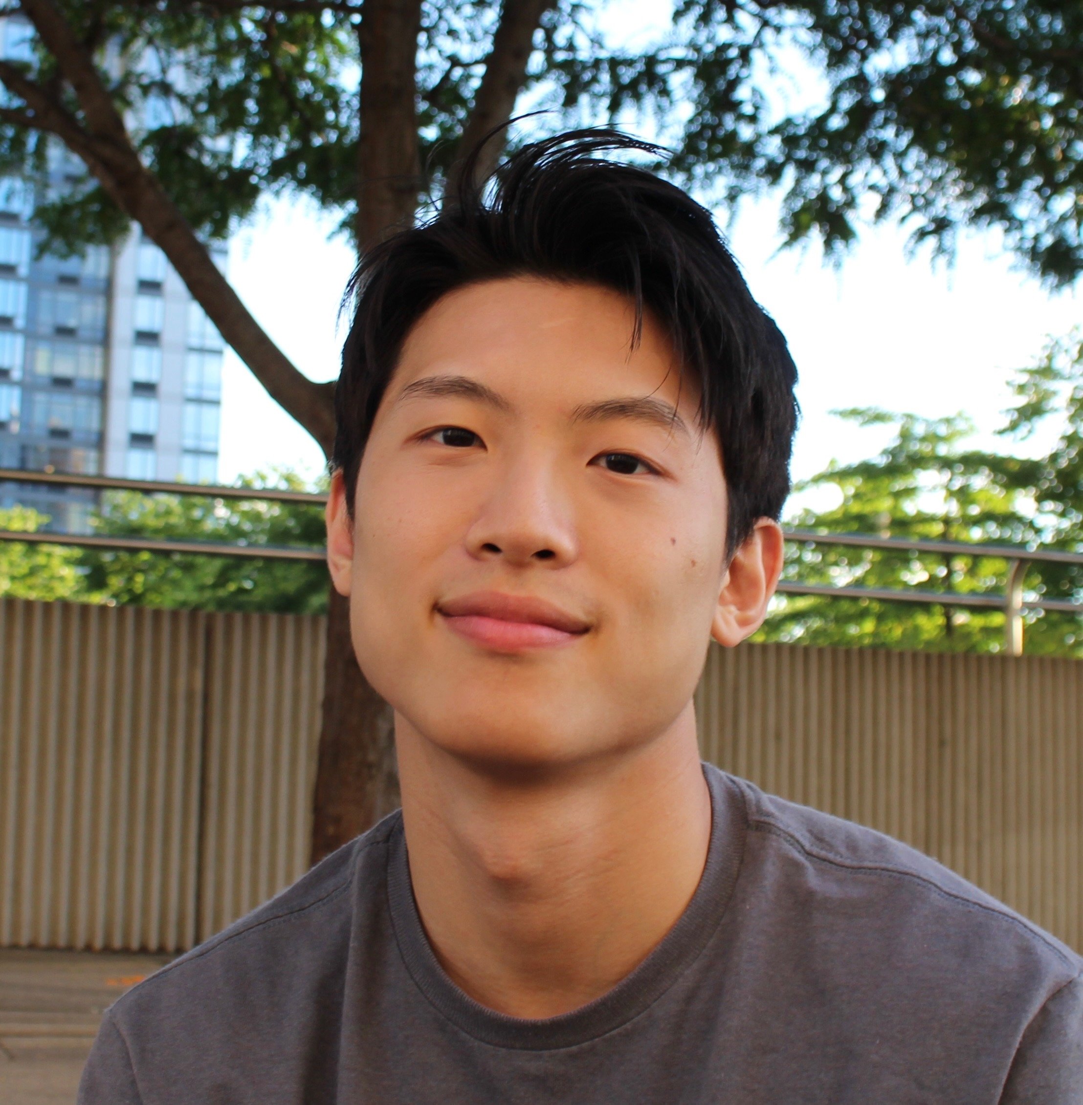 This close-up portrait features a young East Asian man with a calm and friendly demeanor, offering a subtle, almost shy smile directly to the camera. His dark hair is styled with a slight upward tousle, adding a touch of casual charm. He wears a simple dark grey t-shirt, blending into the relaxed outdoor setting. The background, softly blurred, reveals lush green tree foliage and branches, suggesting a park or urban green space. Behind the trees, a modern building with reflective windows hints at a city environment, while a metal railing and corrugated wall complete the urban-natural blend. The overall impression is one of quiet approachability, perhaps caught mid-thought in a peaceful city oasis.