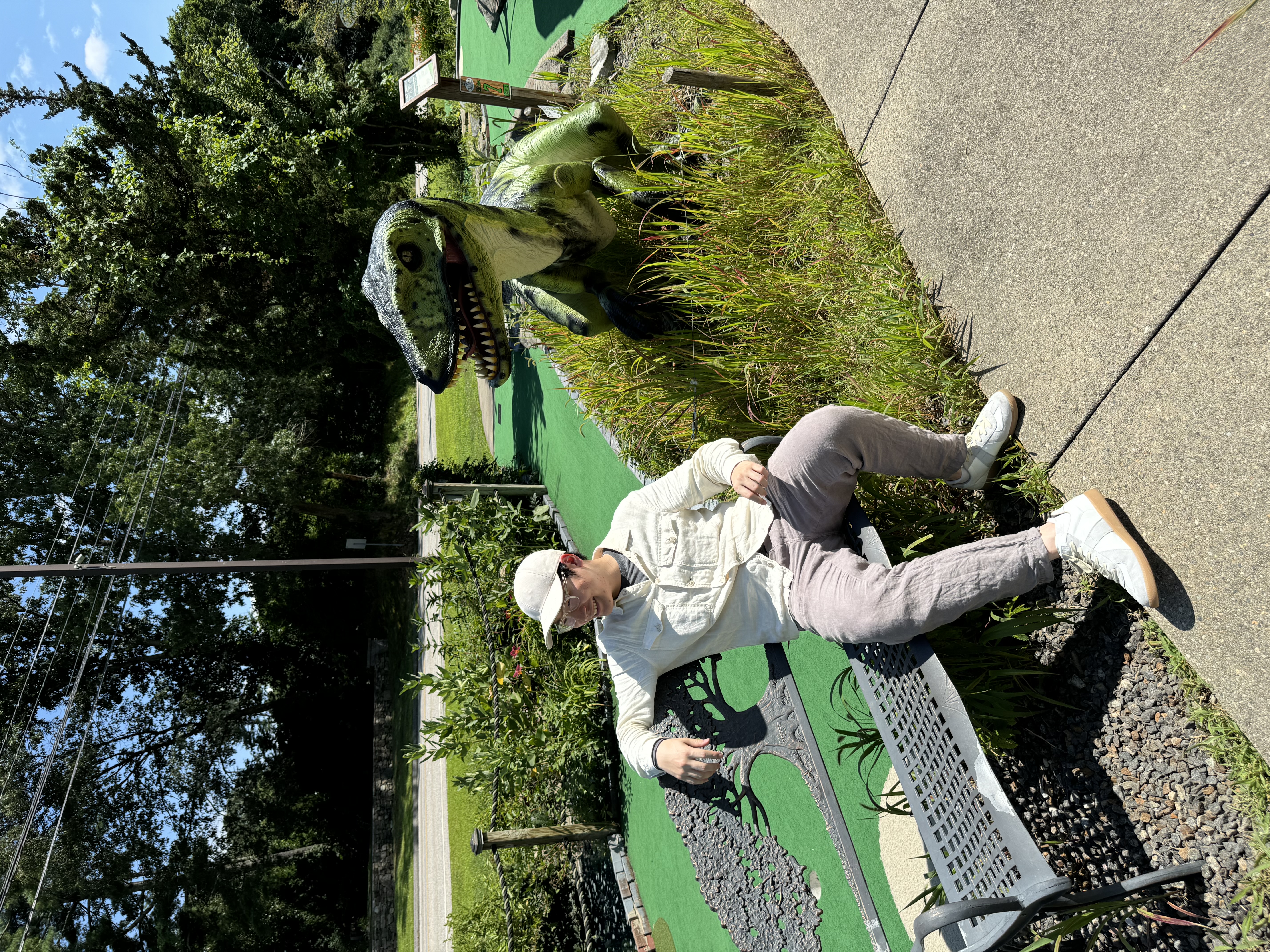 In this playful scene at a mini-golf course, a person is seated on a bench, casually engaging with a smartphone, suggesting a laid-back demeanor. The subject wears a light-colored jacket and a cap, blending in with the bright, sunny atmosphere. Beside them, a whimsical dinosaur figure adds a fun element to the setting. Its vibrant green color and exaggerated features evoke a sense of nostalgia, reminiscent of childhood adventures.

The lush greenery surrounding the mini-golf course enhances the cheerful vibe, while the distant trees and blue sky create a serene backdrop. This playful juxtaposition of the dinosaur and the relaxed human captures the spirit of leisure and enjoyment typical of themed mini-golf venues. Overall, the image exudes a humorous charm, as if the dinosaur is playfully observing the subject's modern-day antics, inviting smiles and laughter.
