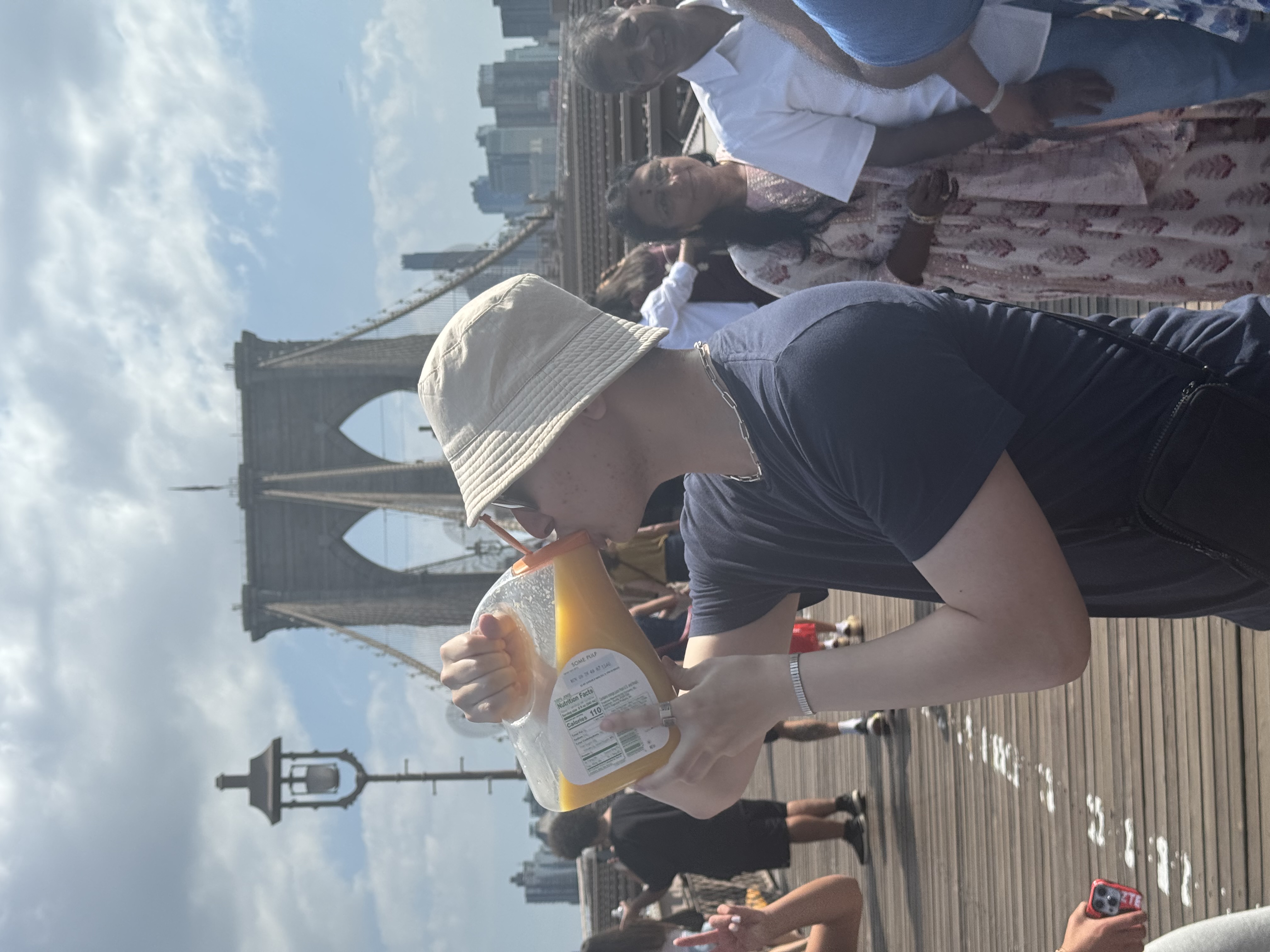 In the image, a person is enjoying a drink from a large container while standing on a wooden pier, with the iconic Brooklyn Bridge prominently in the background. The subject wears a light-colored bucket hat and a dark shirt, suggesting a casual, relaxed demeanor. Their focus is on the drink, creating a moment of enjoyment in a vibrant urban setting.
Surrounding them are a few other people, hinting at a lively atmosphere typical of a day out in New York City. The sky is partly cloudy, adding a soft light to the scene. The Brooklyn Bridge, with its distinctive architecture, serves not only as a beautiful backdrop but also as a symbol of the city’s charm.
This scene captures a lighthearted moment, perhaps humorously emphasizing the contrast between the grandiosity of the bridge and the simplicity of sipping a beverage, making it a delightful snapshot of urban life.