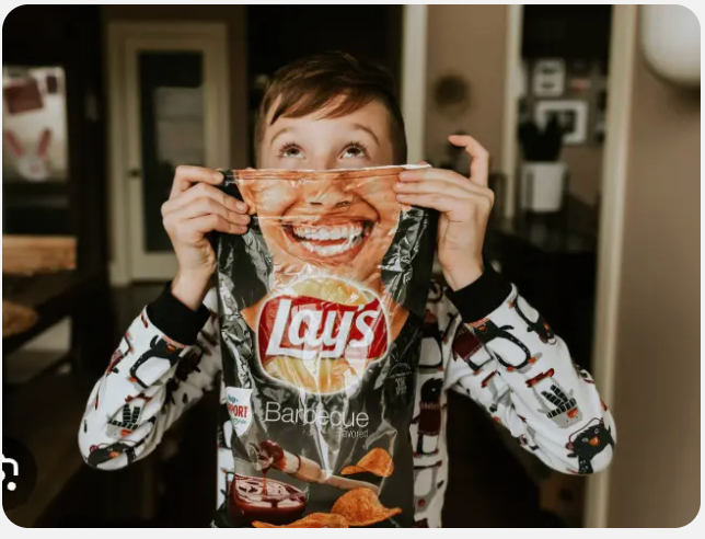 A young child, wearing adorable penguin-patterned pajamas, playfully holds a bag of Lay's Barbecue potato chips up to their face. Their own eyes, wide and looking upwards with a mischievous or curious gaze, peek over the top of the bag, perfectly aligning with the broad, smiling mouth printed on the chip bag. This common playful interaction creates a humorous effect, making it appear as though the chip bag itself has a giant, grinning face with the child's eyes. The background is softly blurred, suggesting a cozy, indoor home environment, which perfectly complements the casual and lighthearted moment captured. The image exudes a sense of innocent fun and childhood joy, highlighting a simple, relatable moment of play.