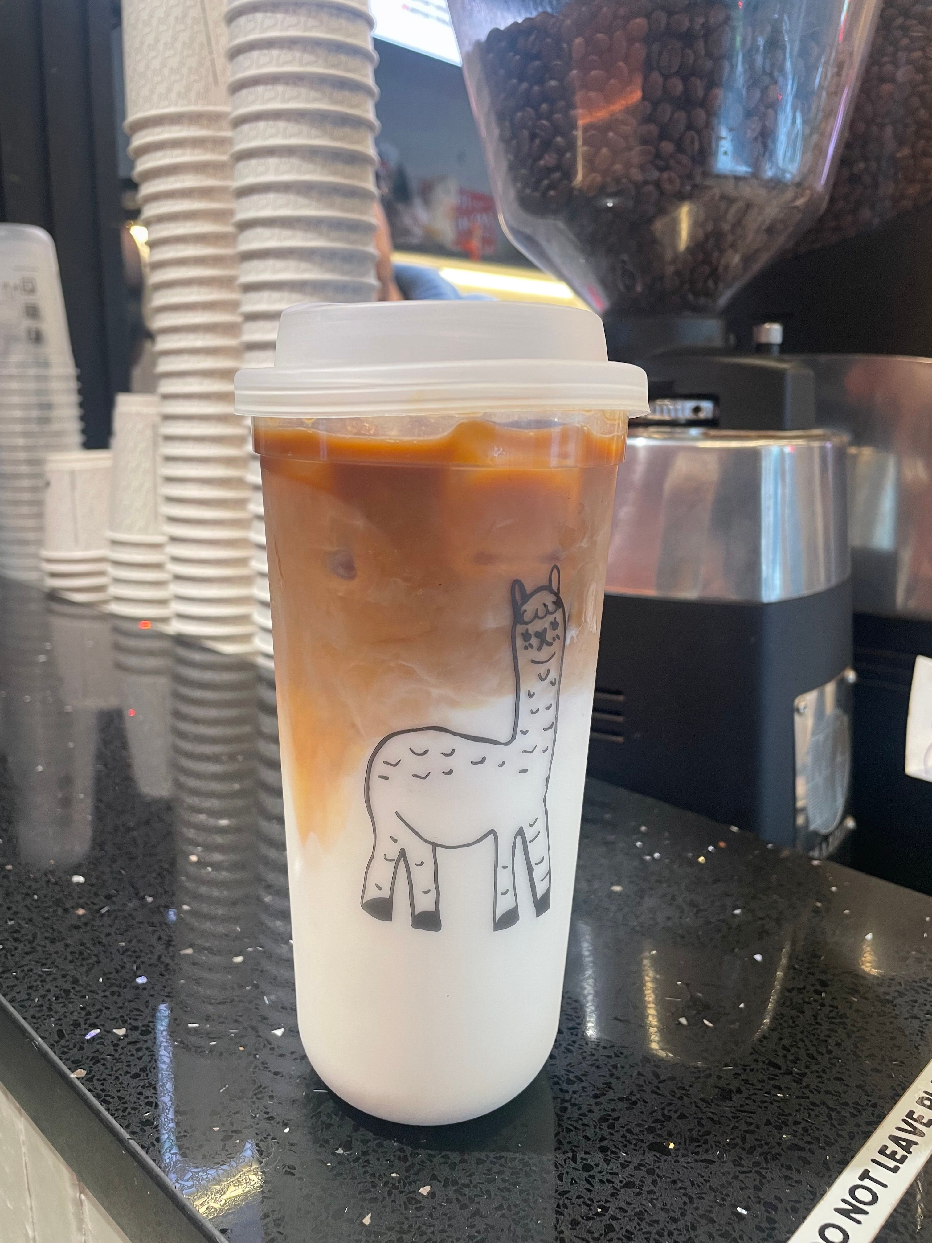 The image features a whimsical coffee cup adorned with a cartoonish llama illustration, prominently displayed against a dark, glossy counter. The drink inside is a delightful mix of iced coffee, with caramel swirling atop a layer of milk, showcasing a playful blend of colors that invite curiosity. The demeanor of the cup might be interpreted as cheerful and inviting, as if it's ready to brighten someone’s day.
In the background, stacks of white paper cups suggest a bustling café atmosphere, hinting at a lively environment filled with coffee enthusiasts. The presence of coffee beans in the grinder adds an earthy aroma to the scene, enhancing the overall experience.
The humorous element lies in the llama, which seems to be enjoying a coffee break itself, as if it’s saying, "I may be a llama, but even I need my caffeine fix!" This quirky touch adds charm to an everyday scenario.