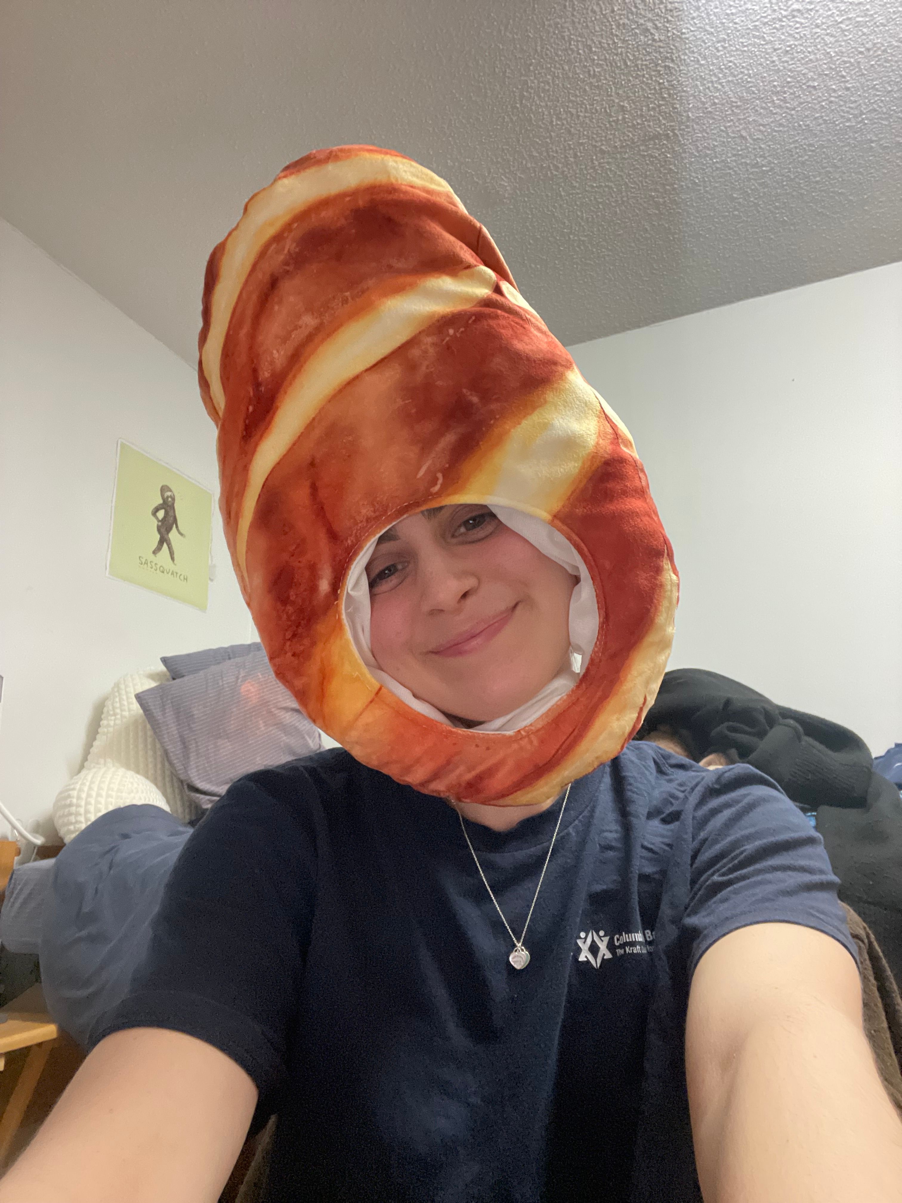A person with a cheerful, playful demeanor grins directly at the camera, sporting an undeniably humorous, oversized novelty bacon hat that frames their face. The hat, resembling a perfectly cooked strip of bacon, dominates the upper half of the image, adding a whimsical touch. In the background, a light-colored wall features a "Sassquatch" poster, a delightful pop culture nod that perfectly complements the subject's quirky headwear. The casual bedroom setting, with pillows visible, suggests a relaxed and fun moment, perhaps a spontaneous selfie. The entire scene exudes a lighthearted, meme-worthy vibe, celebrating humor and individuality.