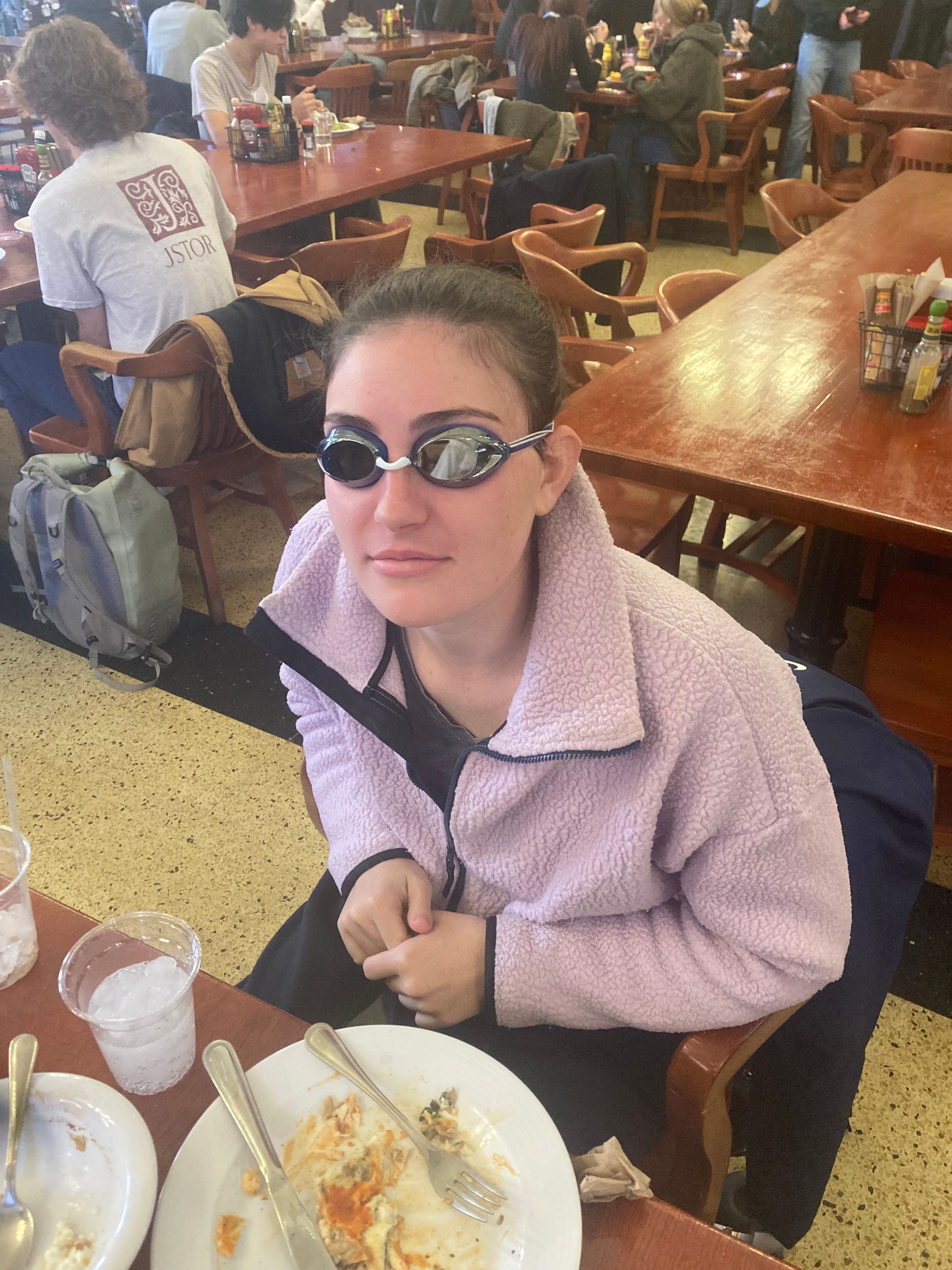 A young woman with dark hair pulled back sits at a wooden table in what appears to be a bustling university dining hall or cafeteria. She wears a light purple fleece jacket and, most notably, a pair of dark-lensed swimming goggles, giving her a comically out-of-place appearance. Her expression is a subtle smirk, looking directly at the camera with a playful demeanor. In front of her are partially eaten plates of food and a cup of ice water, suggesting she's mid-meal. The background is filled with other students or patrons at similar wooden tables, some with backpacks nearby, reinforcing the casual, academic environment. One person in the background wears a t-shirt with a "JSTOR" logo, further hinting at a university setting. The humor lies in the incongruity of her swim goggles in such a mundane, indoor dining context, as if she's ready to dive into her soup.