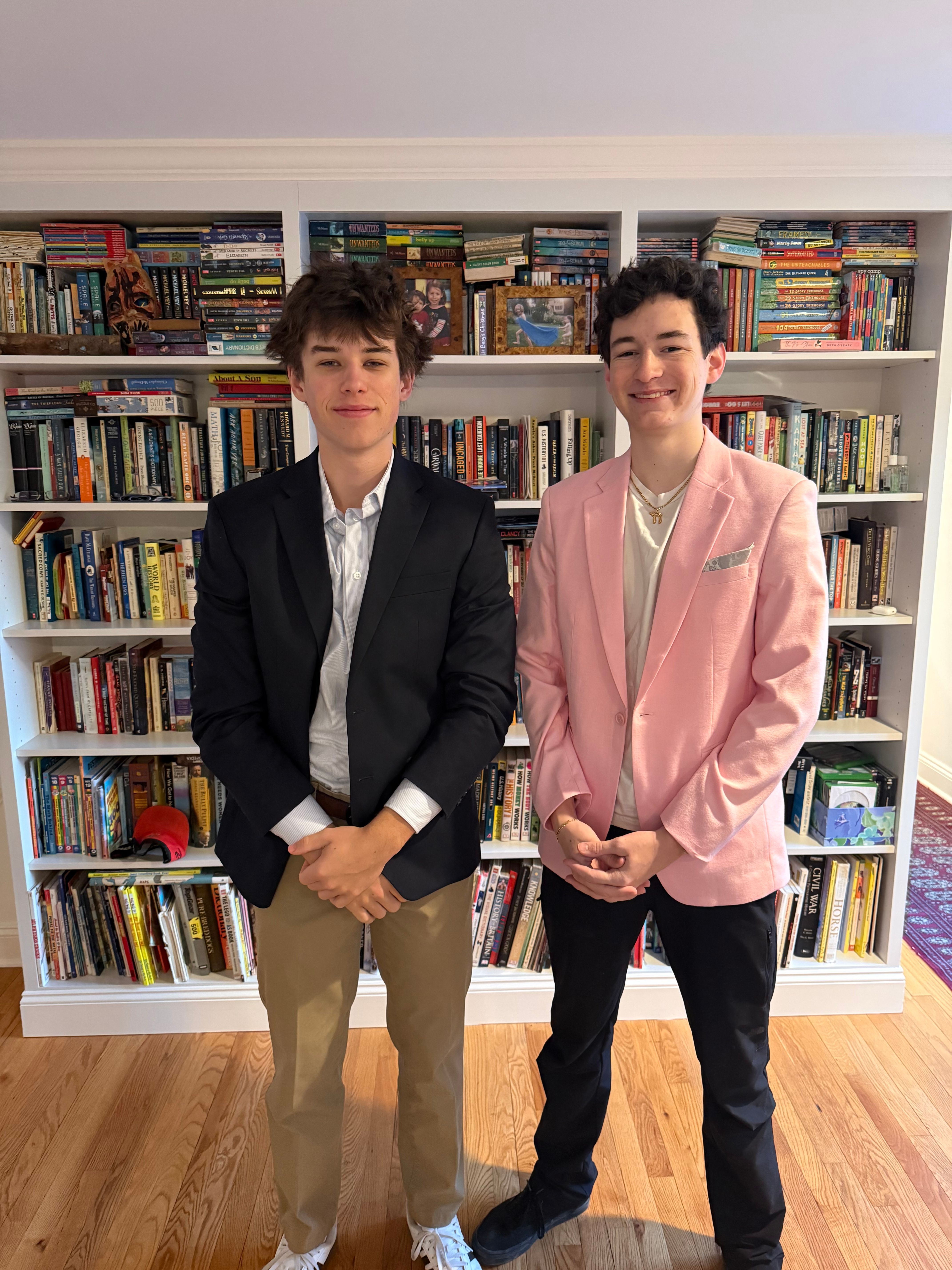 Two young men, possibly brothers, stand smiling confidently in front of a towering, well-stocked white bookshelf. The man on the left sports a dark blazer, striped shirt, khaki pants, and white sneakers, exuding a relaxed charm. His companion on the right beams in a vibrant pink blazer, light t-shirt, black pants, and black sneakers, with a gold pendant adding a touch of flair. Both have their hands clasped, giving them a slightly formal yet approachable demeanor. The background, a vast home library filled with diverse books from 'Warriors' to 'Civil War,' humorously suggests a deep, perhaps eclectic, intellectual foundation or simply a very well-read household. The contrast of their smart blazers with casual sneakers adds a playful, modern twist to their polished appearance.
