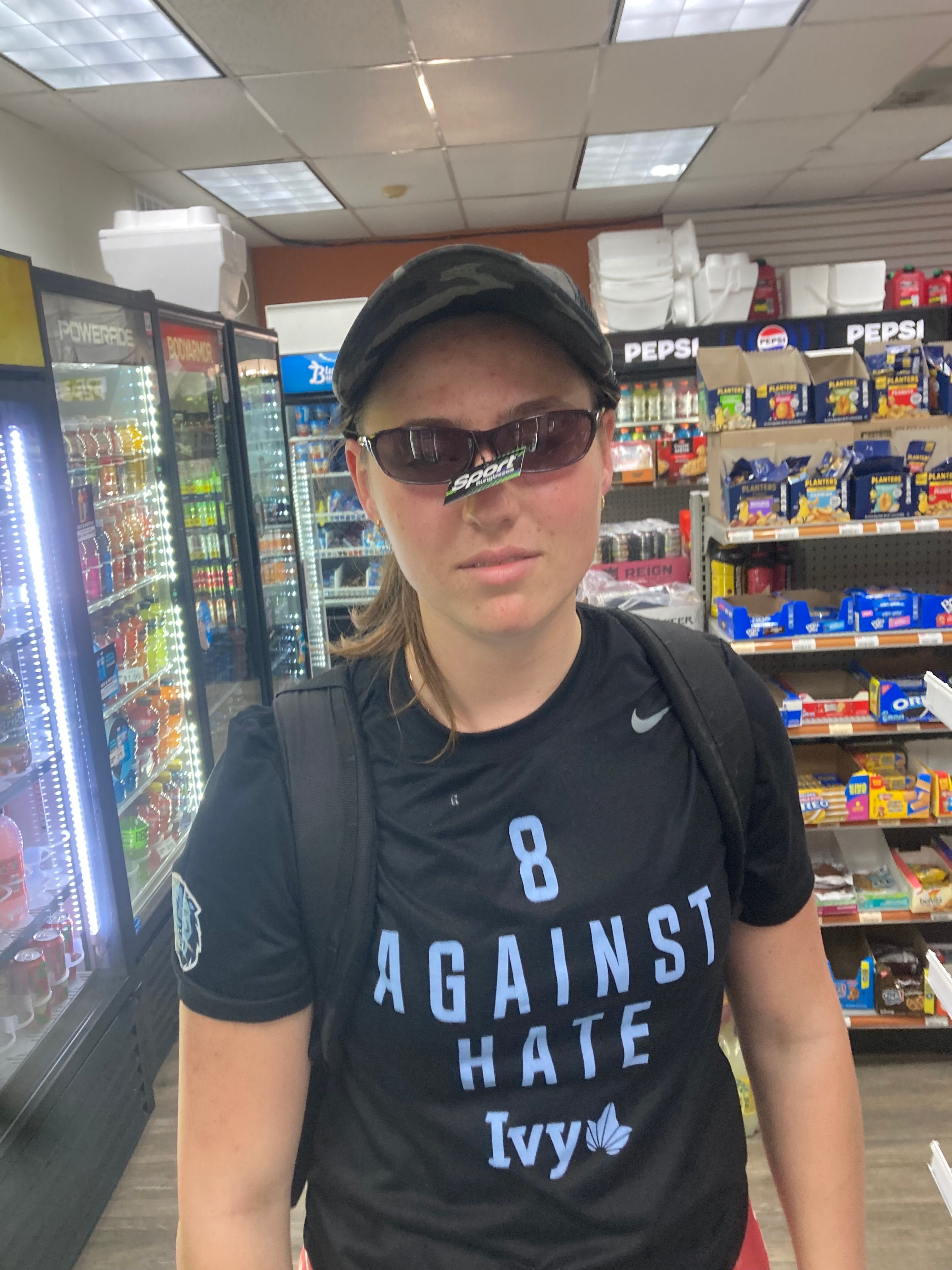 A young woman with a neutral, almost deadpan expression stands in a brightly lit convenience store aisle. She wears a camouflage baseball cap, a black t-shirt with "8 AGAINST HATE Ivy" printed in light blue, and a black backpack. Comically, she's wearing dark 'Sport Sunglasses' indoors, with the green retail tag still dangling from the bridge of her nose, adding a touch of oblivious charm to her demeanor. The background is a typical store setting, featuring beverage coolers stocked with Powerade and Bodyarmor, and shelves displaying popular brands like Pepsi, Planters, and Oreo. The mundane setting contrasts humorously with her sunglasses faux pas, suggesting a quick, unpretentious stop.