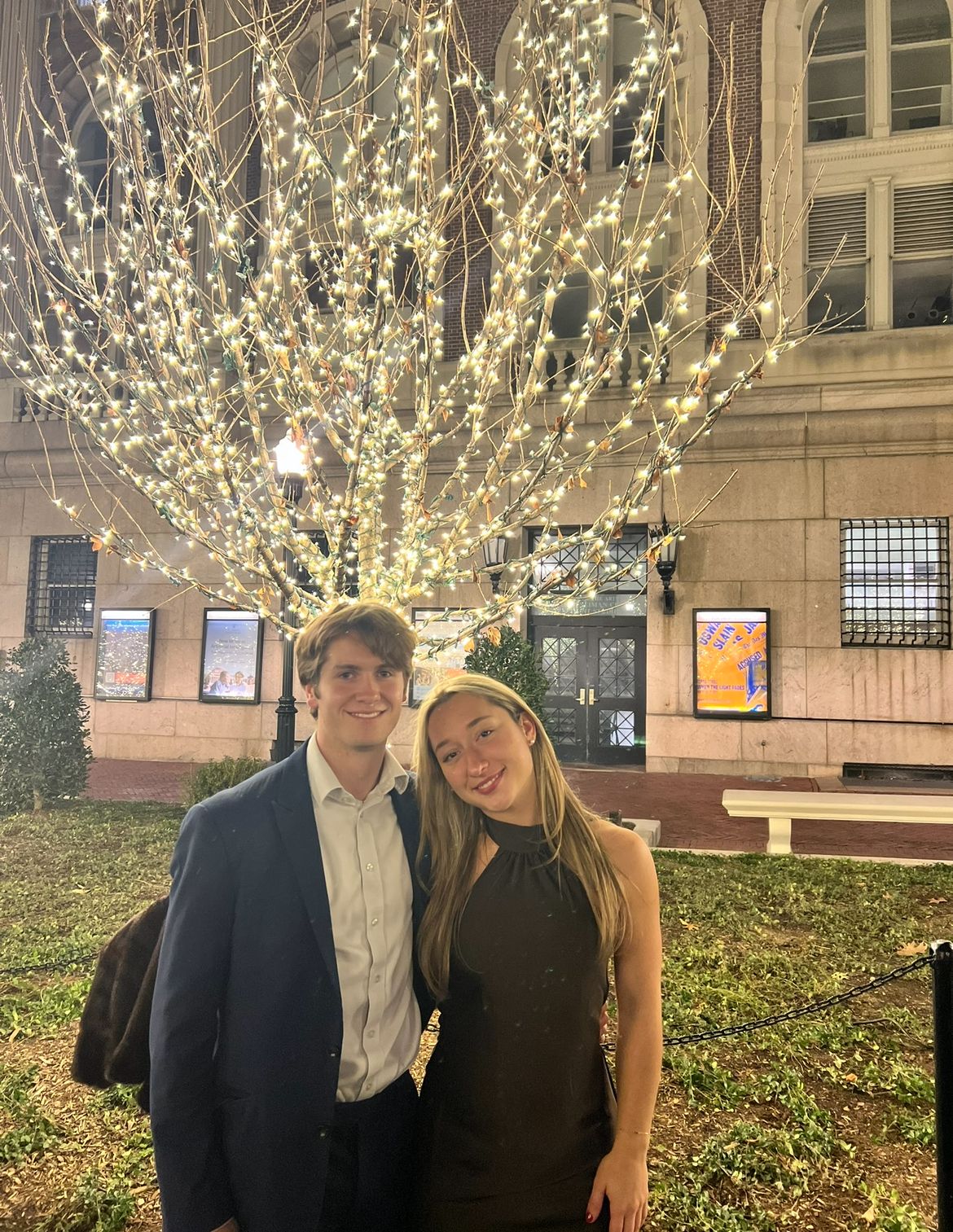 A radiant young couple, dressed for a special occasion, smiles brightly at the camera against a backdrop of a grand, stone university building at night. The man, in a sharp suit jacket, and the woman, in an elegant black halter dress, lean into each other, their joyful expressions suggesting a delightful evening. Behind them, a bare tree is spectacularly, almost comically, adorned with countless warm string lights, creating a dazzling, festive glow that practically screams "holiday card perfection." The building's posters, possibly for a "UVA" event, ground this charming scene in a collegiate context, making one wonder if the tree is trying to steal the spotlight from their undeniable chemistry.