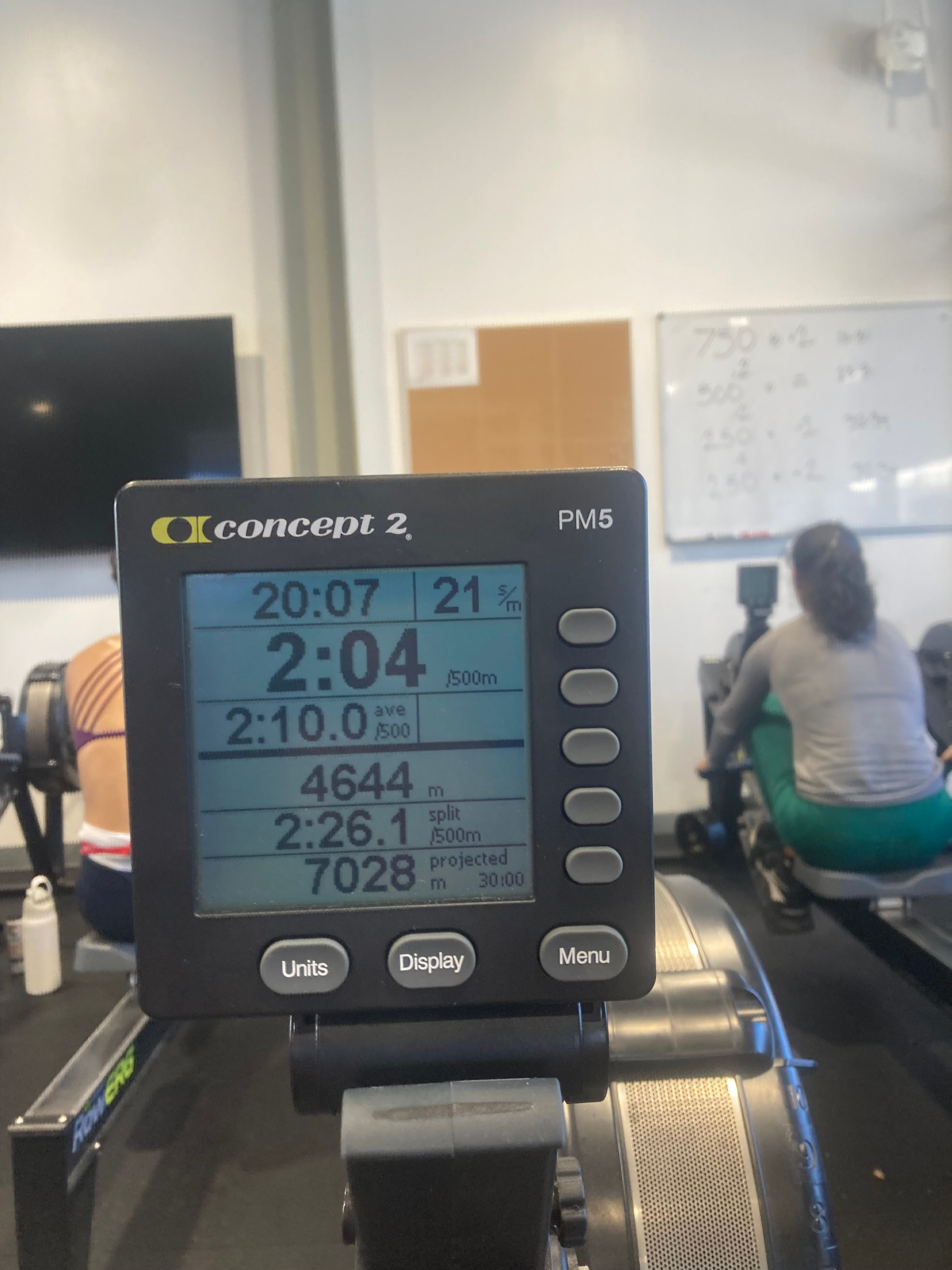 This image provides a close-up, slightly angled view of a Concept2 PM5 rowing machine monitor, acting as the stoic, unblinking eye of fitness. Its digital display, glowing with blue numbers, shows "20:07" elapsed time, "4644 m" distance, and a "2:10.0 /500m" average split, relentlessly tracking the workout. The monitor's "demeanor" is one of unwavering data presentation, perhaps silently judging the rower's pace. In the blurred background, two individuals are seen diligently rowing in what appears to be a gym. One person on the left has distinctive purple athletic tape on their back, while another on the right wears vibrant green leggings. A whiteboard with workout calculations (e.g., "750 * 2") hints at structured training, making the monitor the ultimate scorekeeper in this temple of sweat. The humorous element lies in the monitor's detached, almost robotic, assessment of human exertion.