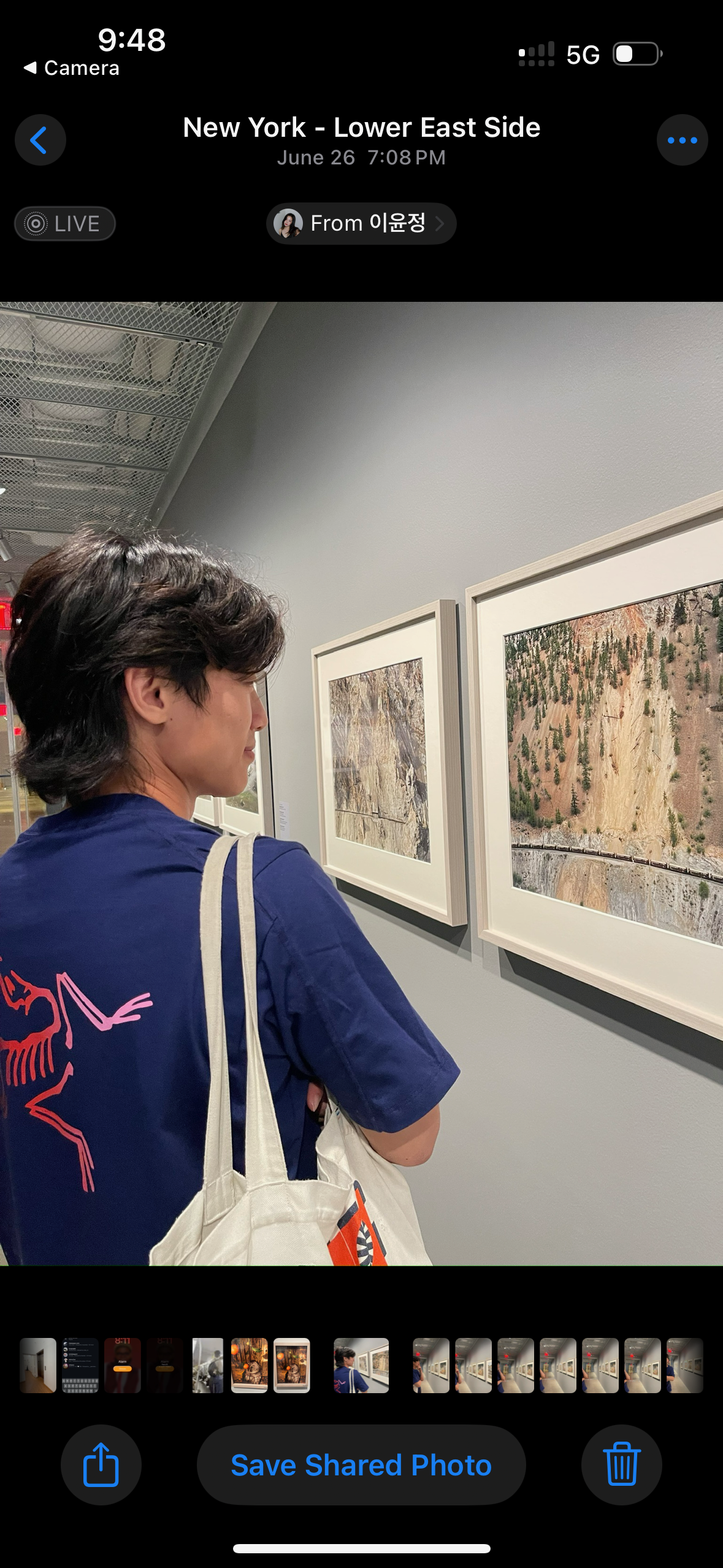 The image features a young person with shoulder-length dark hair, facing a series of framed photographs mounted on a wall. Their demeanor appears contemplative, possibly absorbed in the art displayed. They wear a navy blue shirt adorned with a playful pink skeleton design and carry a light-colored tote bag slung over their shoulder, suggesting a casual, artistic vibe.
The background shows a modern gallery setting, with a sleek, minimalist design that emphasizes the artwork. The photographs seem to depict natural landscapes, adding a serene contrast to the contemporary indoor environment. The lighting is soft, enhancing the overall ambiance of exploration and appreciation for art.
This scene likely captures a moment of reflection in a cultural space, possibly indicating a visit to an exhibit in New York's Lower East Side, known for its vibrant art scene. The playful shirt adds a humorous touch, suggesting the subject is both stylish and open-minded in their artistic pursuits.