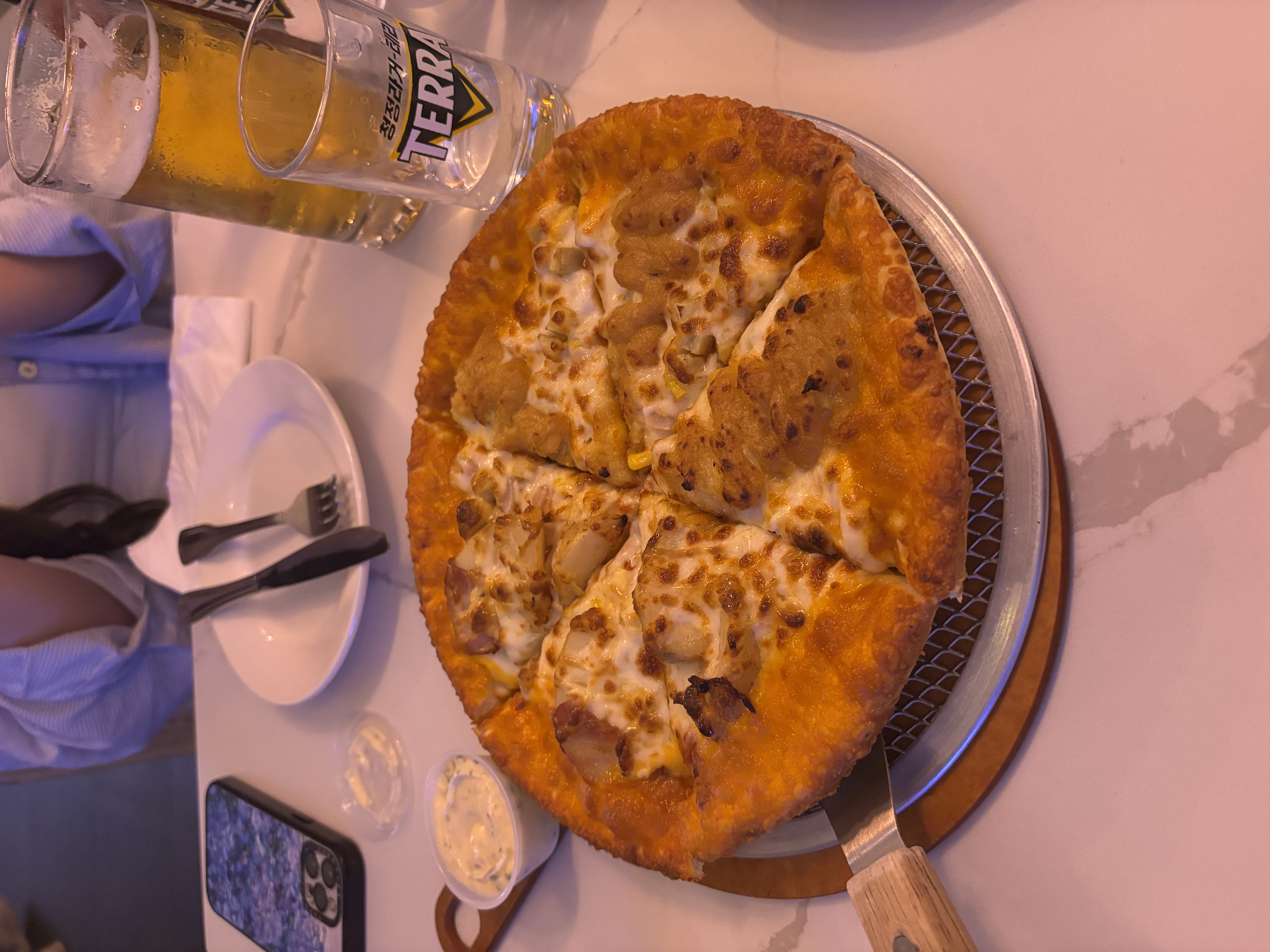 The image showcases a delicious pizza, perfectly golden and cheesy, sitting on a circular metal tray with a wooden handle. The crust appears thick and crispy, suggesting a satisfying crunch with every bite. Nearby, there's a small dish of dipping sauce, likely ranch or garlic, and a clean white plate, fork, and knife set, hinting at a casual dining experience.
In the background, a frosty glass of beer adds to the relaxed atmosphere, suggesting a gathering with friends or a cozy meal out. The dimly lit environment hints at an inviting restaurant setting, perfect for sharing moments over food.
The subject, while not directly visible, seems to be enjoying the meal, as indicated by the hands poised to dig in. The overall vibe radiates warmth and camaraderie, making it a delightful snapshot of culinary enjoyment. Who wouldn't want a slice of that? 🍕