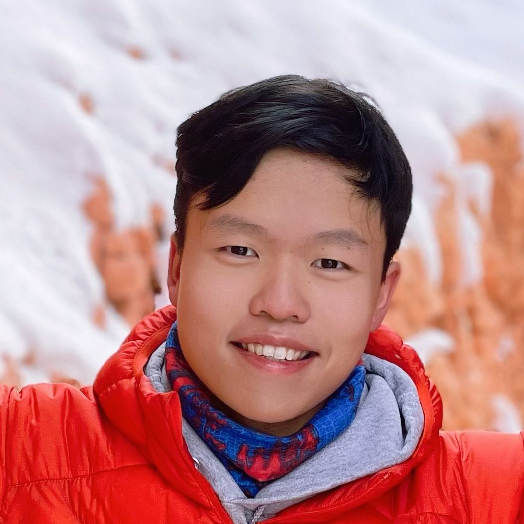 A young man with a radiant, wide smile beams directly at the camera, his eyes sparkling with genuine happiness. He's dressed warmly in a bright red puffer jacket layered over a grey hoodie, with a vibrant blue and red patterned neck gaiter adding a playful touch around his neck. The blurred background suggests a snowy, outdoor environment, possibly a mountain or canyon, with patches of white snow contrasting with hints of orange-brown earth or rock formations. His cheerful demeanor and colorful attire create a humorous contrast with the potentially chilly setting, making him appear utterly delighted to be braving the cold. No famous or recognizable content is found in this image.