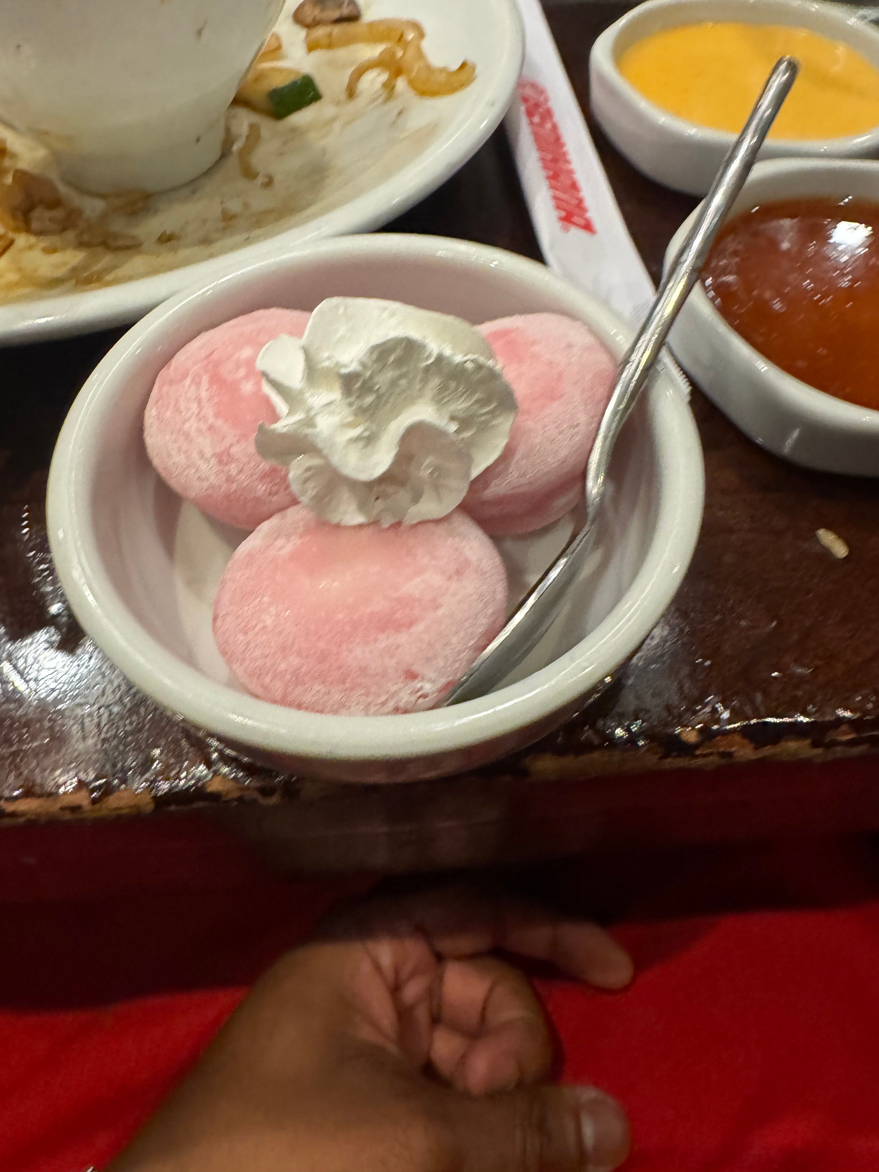 This close-up shot captures a delightful dessert moment, featuring three plump, pink mochi balls nestled in a white bowl. They appear quite bashful, crowned with a fluffy swirl of whipped cream like tiny, elegant hats, while a silver spoon eagerly awaits its turn. In the background, remnants of a savory meal on a plate and small bowls of dipping sauces hint at a satisfying dinner just concluded, setting the stage for this sweet finale. A hand in the foreground seems poised to claim its sugary prize. The scene, likely in a cozy restaurant with a dark wooden table and red tablecloth, exudes a relaxed, post-meal contentment. There are no famous or recognizable elements in this image.