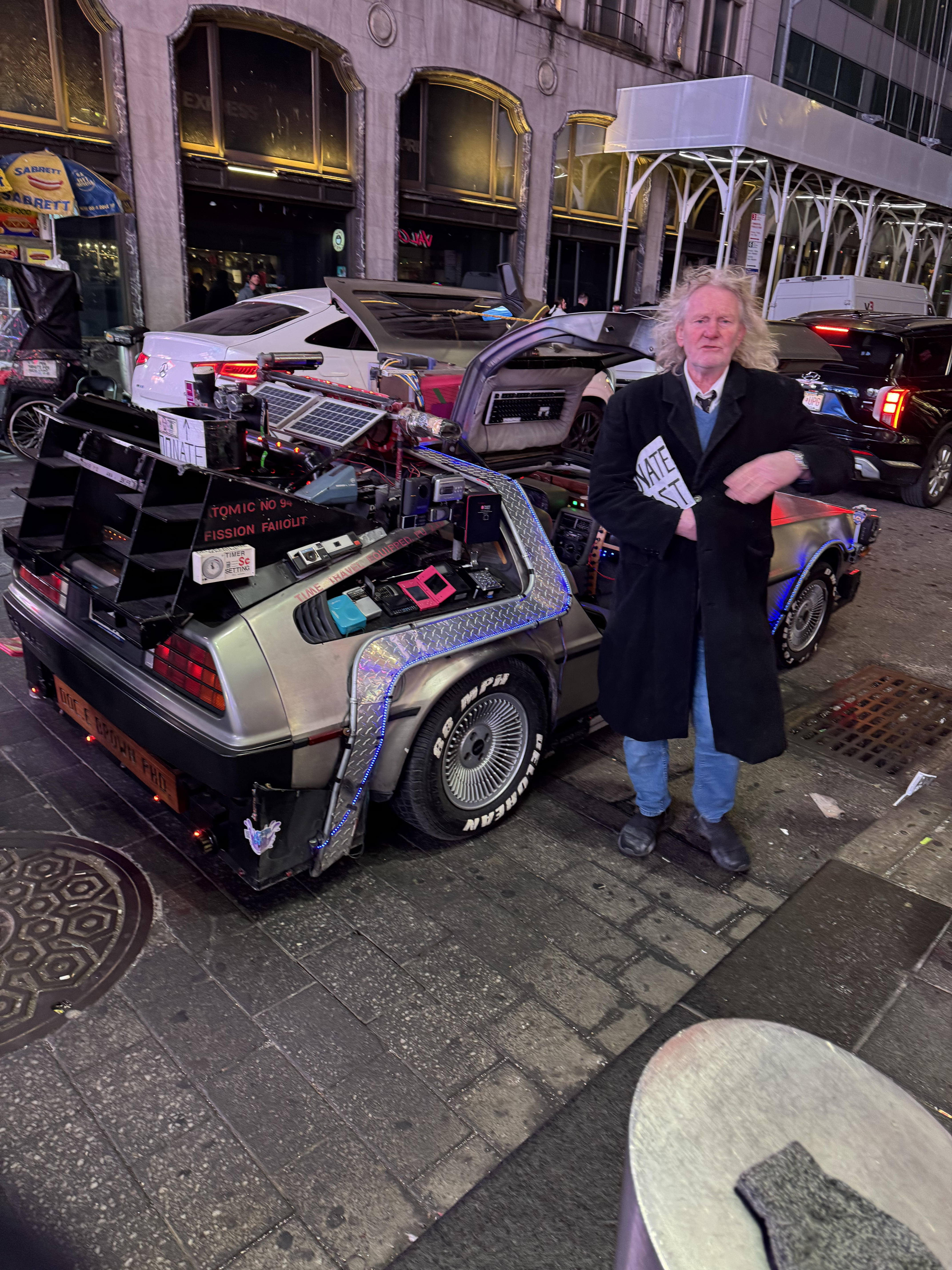In the image, we see a DeLorean time machine, a beloved icon from the "Back to the Future" film series. The vehicle is adorned with various sci-fi gadgets, showcasing its distinctive design. Standing next to it is a man with wild, curly hair and a coat, evoking the character Doc Brown, famously played by Christopher Lloyd. His demeanor appears whimsical, possibly channeling the eccentricity of the character, as he strikes a pose with a playful expression.

The background depicts a bustling urban setting, with cars and city lights, hinting at a lively atmosphere typical of a busy night. The juxtaposition of the futuristic DeLorean against a contemporary cityscape adds a humorous contrast, as if time travel is happening right in the middle of modern life. This scene creates a nostalgic yet amusing reference to the iconic movie, inviting passersby to imagine their own adventures through time.