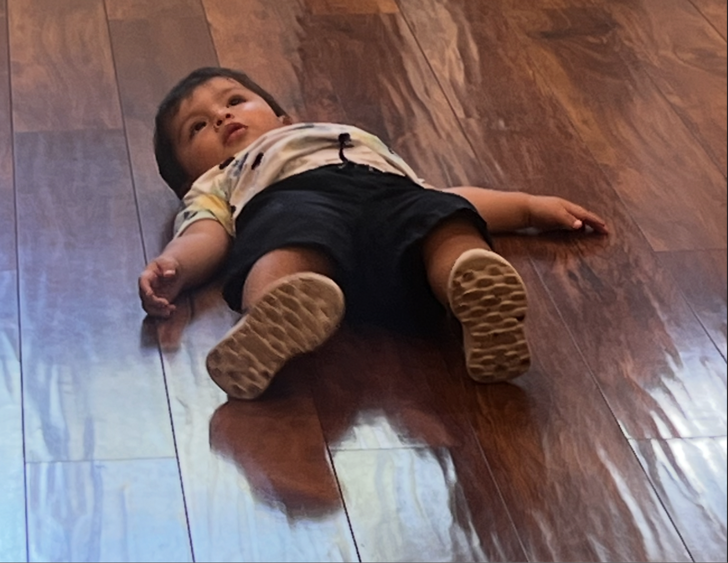 The image depicts a young child lying flat on a polished wooden floor, seemingly in a moment of relaxed contemplation or play. The child’s expression is one of curiosity, gazing up with wide eyes, possibly taking in their surroundings. Dressed in a colorful shirt and black shorts, the child appears comfortable and at ease in this setting.

The warm tones of the wooden floor create a cozy atmosphere, enhancing the innocence and playfulness of the scene. There’s no prominent background detail mentioned, which allows the focus to remain solely on the child’s demeanor and expression. 

Humorously, one might think of this moment as the child declaring a temporary "time-out" from the hustle and bustle of childhood adventures, perhaps pondering the mysteries of life—or simply contemplating their next snack!