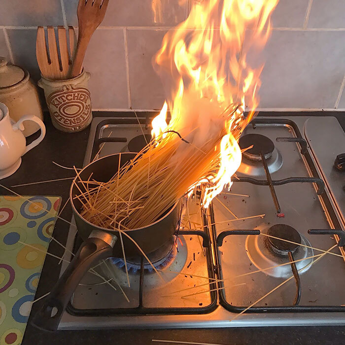 This image captures a dramatic kitchen mishap: a pot of spaghetti on a gas stove, with the dry ends of the pasta bundle engulfed in vibrant orange flames. The spaghetti, seemingly in a fiery protest, stands tall, creating a spectacular, albeit dangerous, culinary inferno. Scattered strands of pasta lie around the burner, casualties of this explosive cooking attempt. The background reveals a typical kitchen counter with a 'KITCHEN UTENSILS' holder, highlighting the domestic setting of this unexpected pyrotechnic display. This scene humorously depicts a cooking disaster of epic proportions, where 'al dente' has been taken to a literal, scorching extreme, making for a truly unforgettable, and likely inedible, meal.
