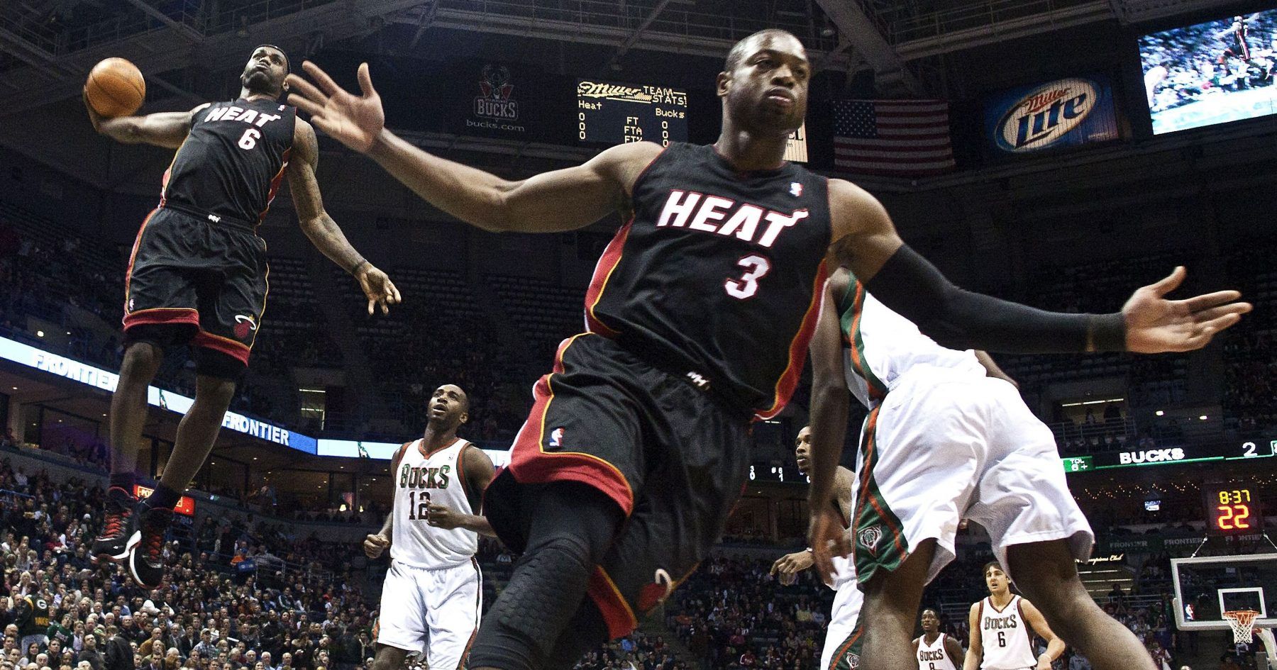 This dynamic NBA image captures Miami Heat stars LeBron James (#6) and Dwyane Wade (#3) in full flight against the Milwaukee Bucks. LeBron, mid-air with the basketball, exhibits intense focus, seemingly defying gravity as he prepares to score. His demeanor is one of powerful determination. Beside him, Wade, with arms dramatically outstretched and a serious expression, appears to be orchestrating the play or reacting to the unfolding action, almost comically trying to encompass the entire court with his presence. The background, filled with cheering fans and arena signage like "Miller Lite" and "Bucks," firmly places this high-stakes moment within a professional basketball game. The Bucks players, looking up, seem caught in the Heat's aerial ballet, highlighting the dominant athleticism on display.