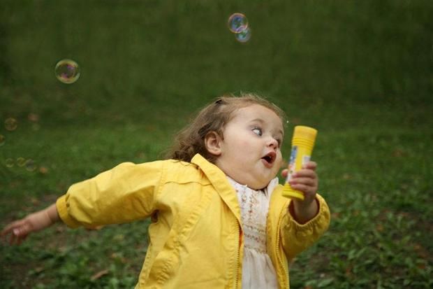 This iconic image features Chloe Clem, famously known as the "Surprised Chloe" meme, captured in a moment of pure, wide-eyed astonishment. Dressed in a vibrant yellow jacket over a white dress, she holds a yellow bubble wand, her mouth agape and eyes fixed upwards and to the right, tracking a floating bubble. Her expression perfectly conveys a mix of wonder and slight bewilderment, as if the bubbles are performing a magic trick just for her. The blurred green background of grass or foliage places her outdoors, a natural setting for playful bubble blowing, enhancing the innocent, whimsical context. The humor lies in her exaggerated, almost theatrical reaction to something as simple as bubbles, making her a relatable symbol for unexpected delights or mild confusion.