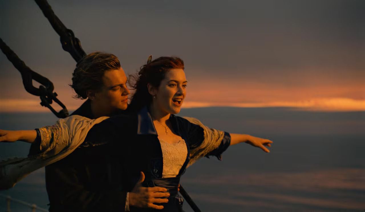 This iconic image captures Leonardo DiCaprio as Jack and Kate Winslet as Rose in the famous "I'm flying!" scene from the movie Titanic. Rose stands at the ship's bow, arms outstretched, her face beaming with pure exhilaration and joy, mouth open in a shout of delight. Jack stands behind her, holding her waist, gazing at her with a tender, admiring expression, providing a sense of security and shared wonder. The golden-orange hues of the sunset paint the sky and reflect on the vast ocean behind them, perfectly setting a romantic and adventurous mood. The ship's rigging is visible on the left, grounding the scene on the grand vessel. This moment perfectly encapsulates their burgeoning love and freedom, a fleeting, blissful escape before the inevitable. The humor lies in the sheer, unbridled, almost childlike joy of Rose, completely lost in the moment, oblivious to the impending doom.
