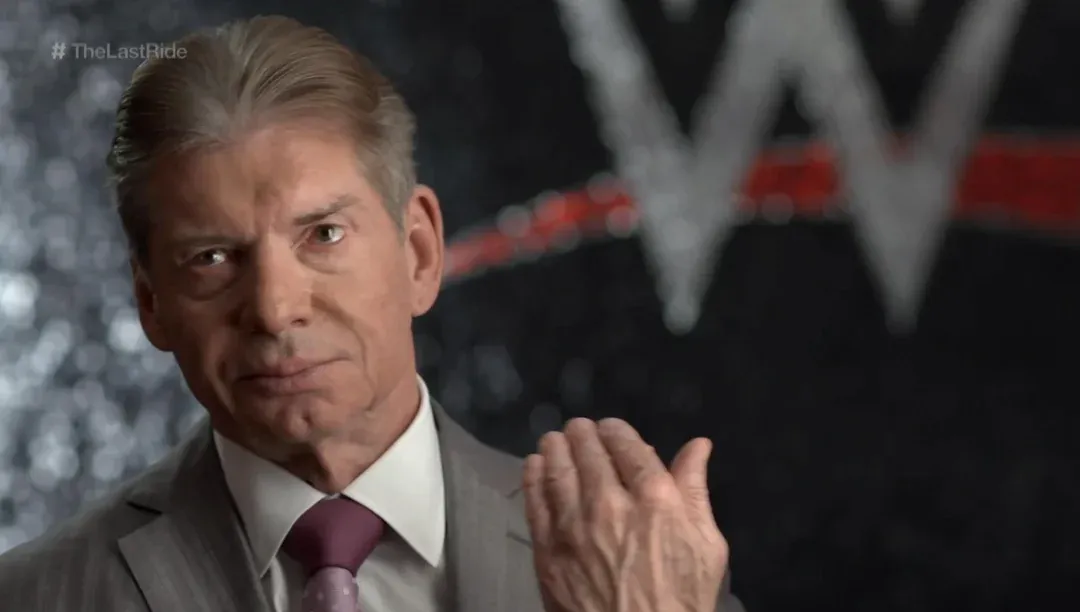 Vince McMahon, the iconic WWE chairman, stares intensely into the camera with a stern, unyielding expression, his brow slightly furrowed. Dressed in a sharp grey suit and a purple tie, his right hand is raised, palm open, as if delivering a definitive, no-nonsense statement. The background features a blurred but recognizable WWE logo, with the hashtag "#TheLastRide" prominently displayed in the top left. This image captures McMahon in a serious moment, likely from the documentary series about The Undertaker, where he's known for his dramatic pronouncements. His gaze suggests he's either about to drop a bombshell or is simply contemplating the next outrageous storyline, perhaps wondering if he can still pull off a "You're Fired!" with the same gravitas.
