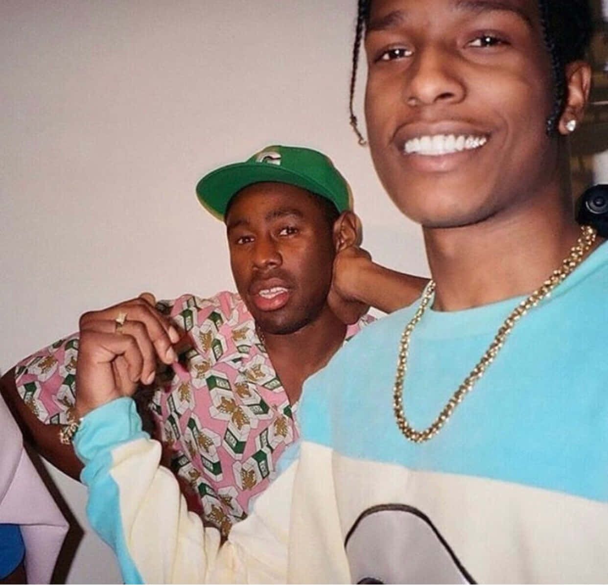 This candid photo captures a humorous moment between musicians A$AP Rocky and Tyler, The Creator. A$AP Rocky, in the foreground, beams with a wide, confident smile, showcasing his bright white teeth and a gold chain over his light blue and white top. Behind him, Tyler, The Creator, wearing a green cap and a pink money-patterned shirt, sports a comically surprised or slightly bewildered expression with his mouth agape, his hand resting casually on A$AP Rocky's shoulder. The plain, light background emphasizes their playful interaction, suggesting an informal, backstage vibe. It's a classic snapshot of their dynamic friendship, full of personality and contrasting energies.