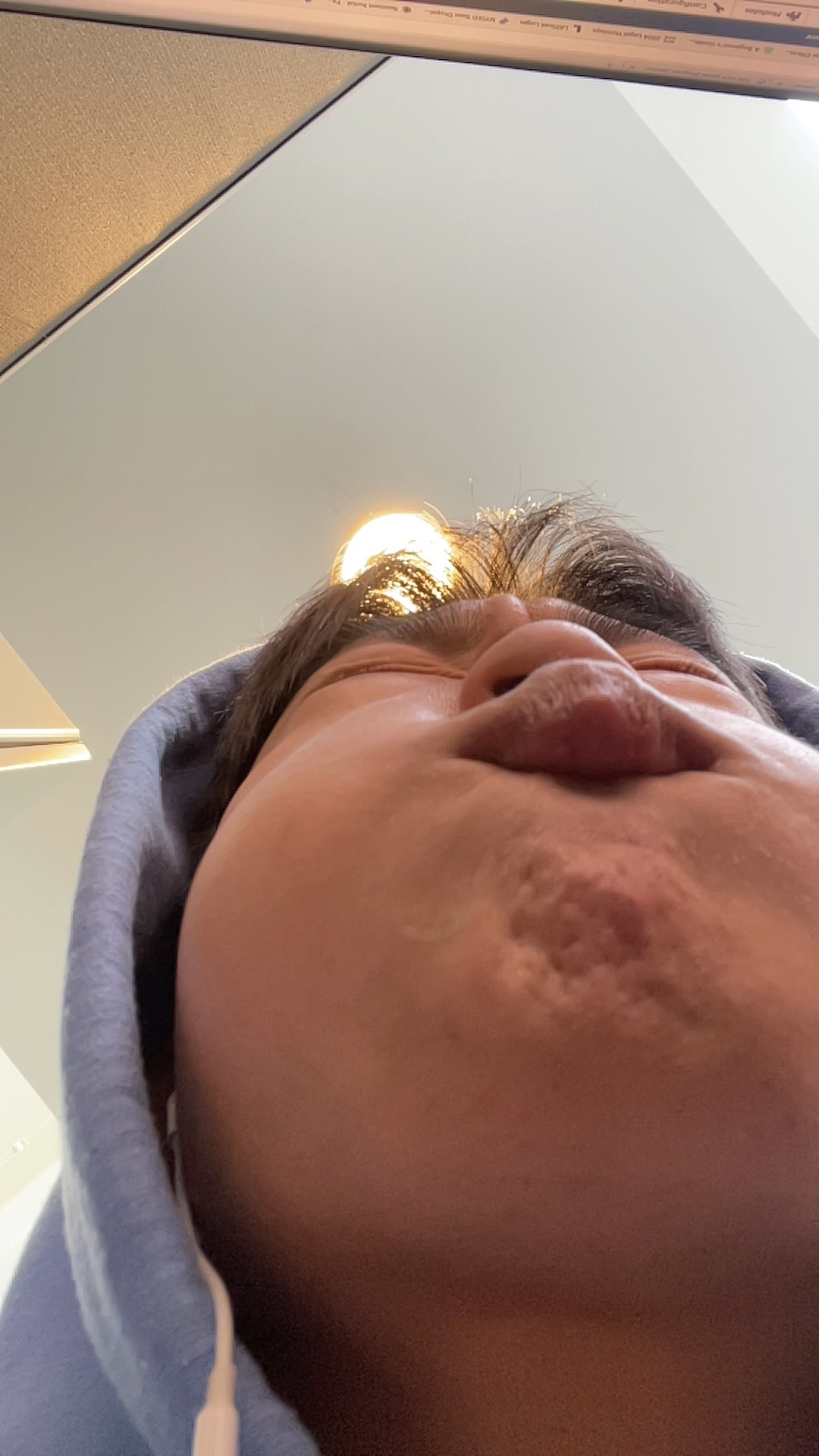 The image depicts a close-up of a person's face, captured from a low angle. The subject appears to be making a funny or exaggerated expression, with their cheeks puffed out and their lips pursed. This creates a humorous and somewhat playful demeanor, as if they are either trying to blow a bubble or making a silly face for the camera.
The background is minimalistic, featuring soft colors and lighting that suggests an indoor setting, possibly a room with a ceiling light visible in the top center of the frame. The soft ambiance complements the lightheartedness of the subject’s expression.
Overall, the image evokes a sense of fun and spontaneity, reminiscent of playful moments shared with friends or family, capturing the joys of casual photography. It's a classic example of the kind of candid moments that often lead to laughter and smiles.