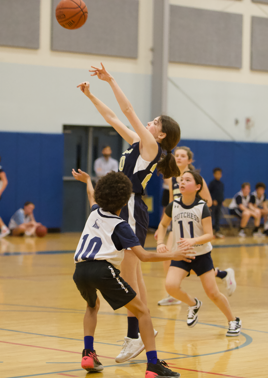 In this dynamic basketball scene, a young girl in a dark jersey (number 0) is captured mid-jump, releasing a basketball towards the hoop. Her expression shows intense focus and determination, embodying the energy of the game. The player in the white jersey (number 10) appears to be attempting to block the shot, with a look of concentration mixed with surprise. Other players in the background, including a girl in a white jersey (number 11), are also engaged in the action, highlighting the competitive atmosphere of the match.
The background features a gymnasium setting, with blue walls and spectators visible, enhancing the context of a spirited youth sports event. The hardwood floor and the placement of players suggest a well-organized game, typical of school-level competitions. This image perfectly captures the enthusiasm and spirited competition of youth basketball, showcasing not just the sport but also the camaraderie among the players.
