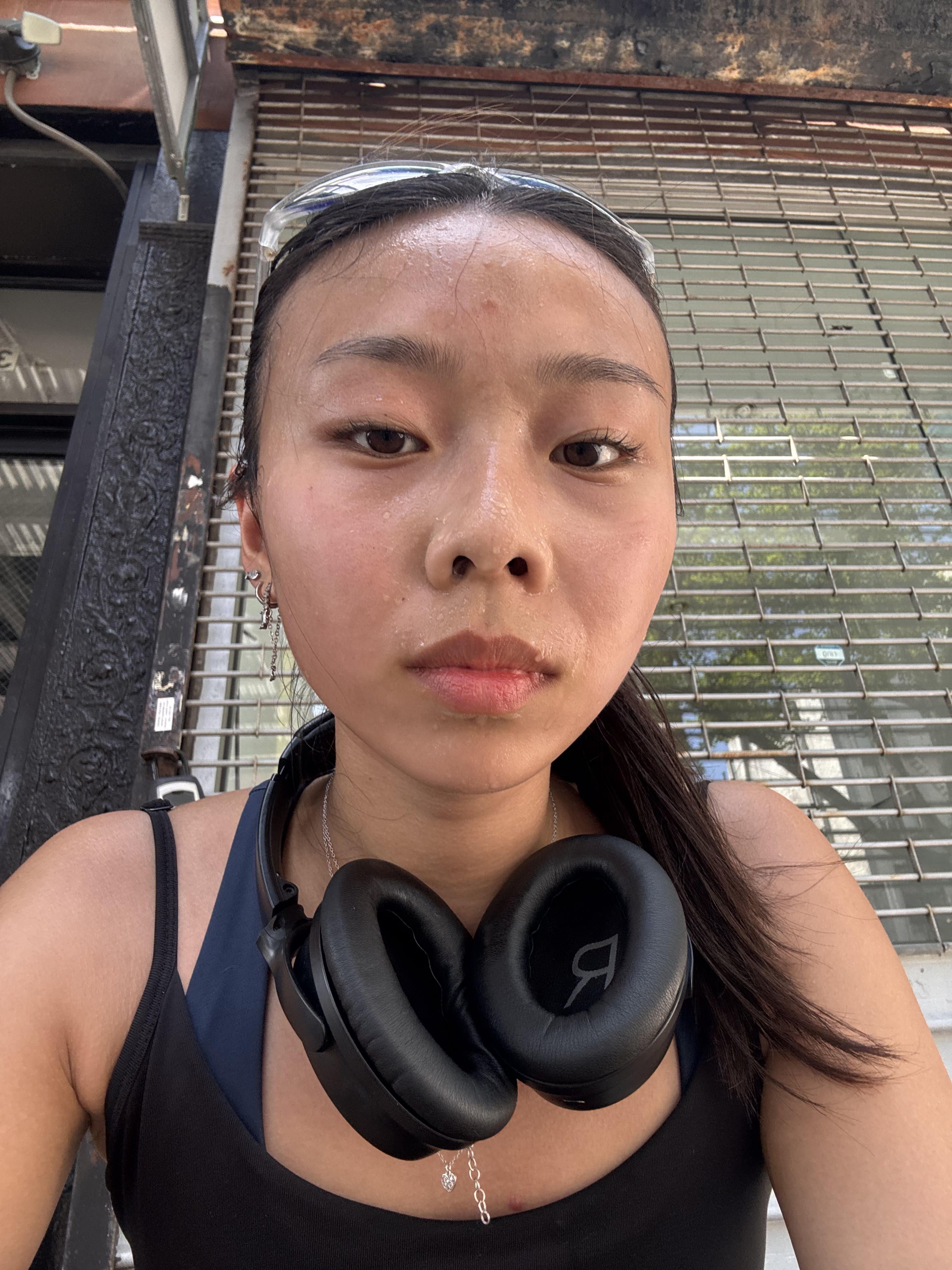 This close-up selfie captures a young woman, visibly glistening with sweat, suggesting a recent intense workout or a very hot day. Her demeanor is neutral, perhaps a mix of exhaustion and satisfaction, as she gazes directly at the camera. She wears clear-framed sunglasses pushed up on her head and large black over-ear headphones resting around her neck, indicating she's likely just finished exercising or is taking a break. Her dark hair is pulled back, and she's dressed in a black tank top over a blue sports bra. The background features a grimy, rusted metal roll-down security gate, typical of an urban storefront, with glimpses of green foliage peeking through. This gritty urban backdrop humorously highlights her post-exertion state, a true warrior of the city streets, unbothered by her sweaty glow.