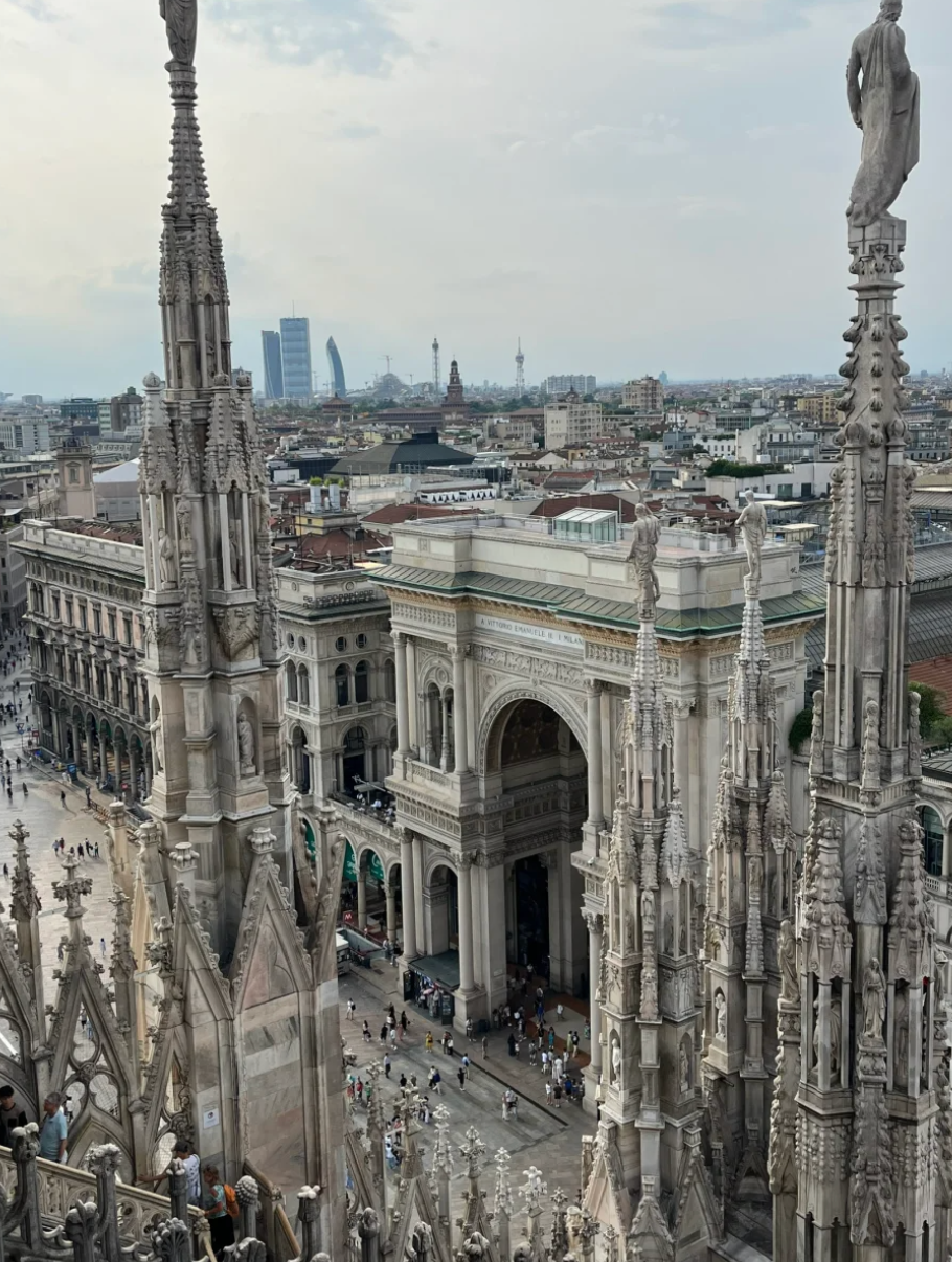 From the majestic rooftop of the Duomo di Milano, a breathtaking panorama unfolds. The intricate Gothic spires, adorned with countless statues, stand like ancient sentinels, their weathered stone faces stoically observing the bustling Piazza del Duomo below. One particular statue, perched high, seems to be pondering the tiny ant-like humans scurrying into the grand archway of the Galleria Vittorio Emanuele II, perhaps wondering if they're all late for an espresso. In the distance, modern skyscrapers playfully poke through the hazy sky, a humorous contrast to the centuries of history beneath. The entire scene, under an overcast sky, captures Milan's timeless blend of grandeur and contemporary life, a silent testament to enduring beauty.