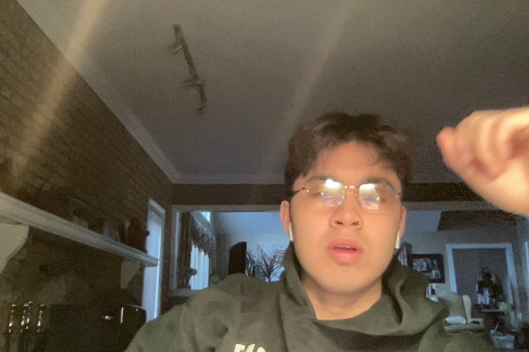 This dimly lit image captures a young man mid-expression, wearing glasses and a dark hoodie with earbuds. His mouth is slightly agape, and his eyes are wide, giving him a look of mild surprise or perhaps deep contemplation, as if he's just heard something utterly baffling. The reflections in his glasses add to his slightly bewildered charm. A blurry hand, possibly his own, intrudes from the top right, adding a touch of spontaneous chaos. The background reveals a cozy, if somewhat dark, home interior with a brick-textured wall, a white shelf, and glimpses into other rooms. The low light and candid nature make it feel like a screenshot from a late-night video call, perfectly capturing that 'wait, what?' moment. It's a relatable snapshot of everyday digital life.