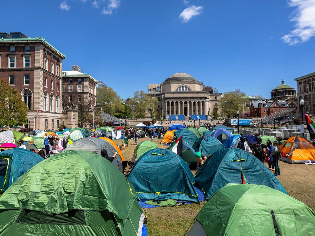 This image depicts a lively student protest encampment, strongly resembling the Columbia University protests, under a brilliant blue sky. A colorful array of tents – greens, oranges, blues – blankets the university lawn, creating a temporary, determined village. These tents, like a flock of brightly plumed, slightly rumpled migratory birds, have landed with a collective demeanor of steadfast resolve. Students move purposefully among them, fostering a vibrant, if unconventional, community. In the background, the grand, classical university buildings, with their stoic stone facades, appear to be silently observing, perhaps muttering about this unexpected outdoor dormitory. The juxtaposition of the makeshift camp against the venerable architecture creates a humorous tension, highlighting a significant cultural event where temporary structures challenge established institutions. It's a campus-wide slumber party with a serious message.