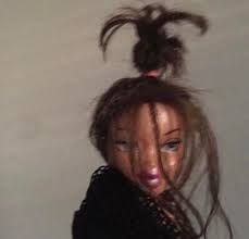 The image captures a close-up of a Bratz doll's head, showcasing a hilariously disheveled yet undeniably sassy demeanor. Its long, brown hair is in utter chaos, with a wild, spiky bun defying gravity on top and strands falling haphazardly across its face, partially obscuring one of its signature large, blue eyes. The doll's expression, with half-lidded eyes and slightly pursed, glossy pink lips, conveys a humorous "just rolled out of bed" or "I woke up like this" attitude, contrasting sharply with the typically glamorous Bratz aesthetic. The plain, light-colored background emphasizes the doll's unkempt yet confident presence, making it a relatable and amusing depiction of a bad hair day, anthropomorphizing the doll's struggle with morning hair.