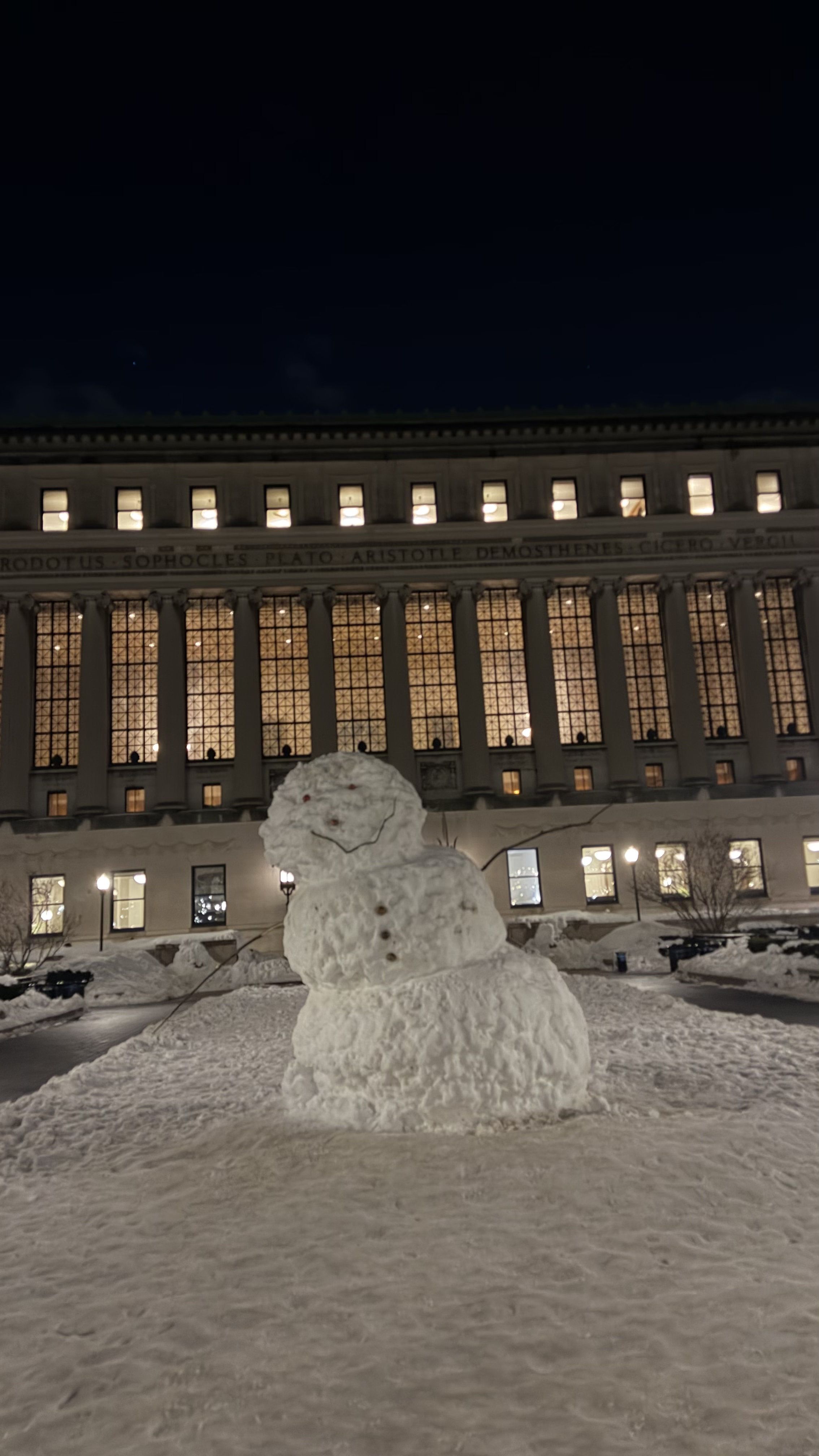 A jovial, three-tiered snowman stands proudly on a snow-covered path at night, its twig-smile slightly askew, giving it a charmingly mischievous demeanor. Its small, red eyes seem to twinkle with frosty delight, perhaps contemplating the wisdom emanating from the grand building behind it. This imposing structure, clearly Columbia University's Butler Library, is brightly lit, revealing the names of ancient scholars like Plato and Aristotle etched above its windows. The snowman, a temporary student of the classics, seems to be enjoying a quiet, snowy evening, perhaps pondering the profound thoughts of its intellectual neighbors. The scene is a humorous blend of academic gravitas and wintery whimsy.