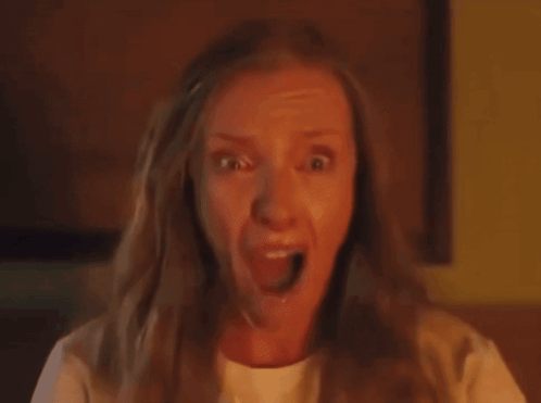 This close-up shot captures Toni Collette in a moment of extreme terror, her face contorted in a wide-mouthed scream, eyes bulging with fear. The intense orange lighting bathes her features, highlighting sweat or tears, and suggests a dramatic, possibly fiery, event unfolding. This iconic image is from the horror film 'Hereditary,' where Collette's character, Annie Graham, experiences profound psychological and supernatural distress. The background is dark and indistinct, emphasizing her raw emotion, but the warm glow implies an ominous, internal setting, perfectly aligning with the film's terrifying atmosphere. Her expression is so visceral it's almost comically relatable to anyone facing an unexpected jump scare or realizing they forgot to turn off the stove.