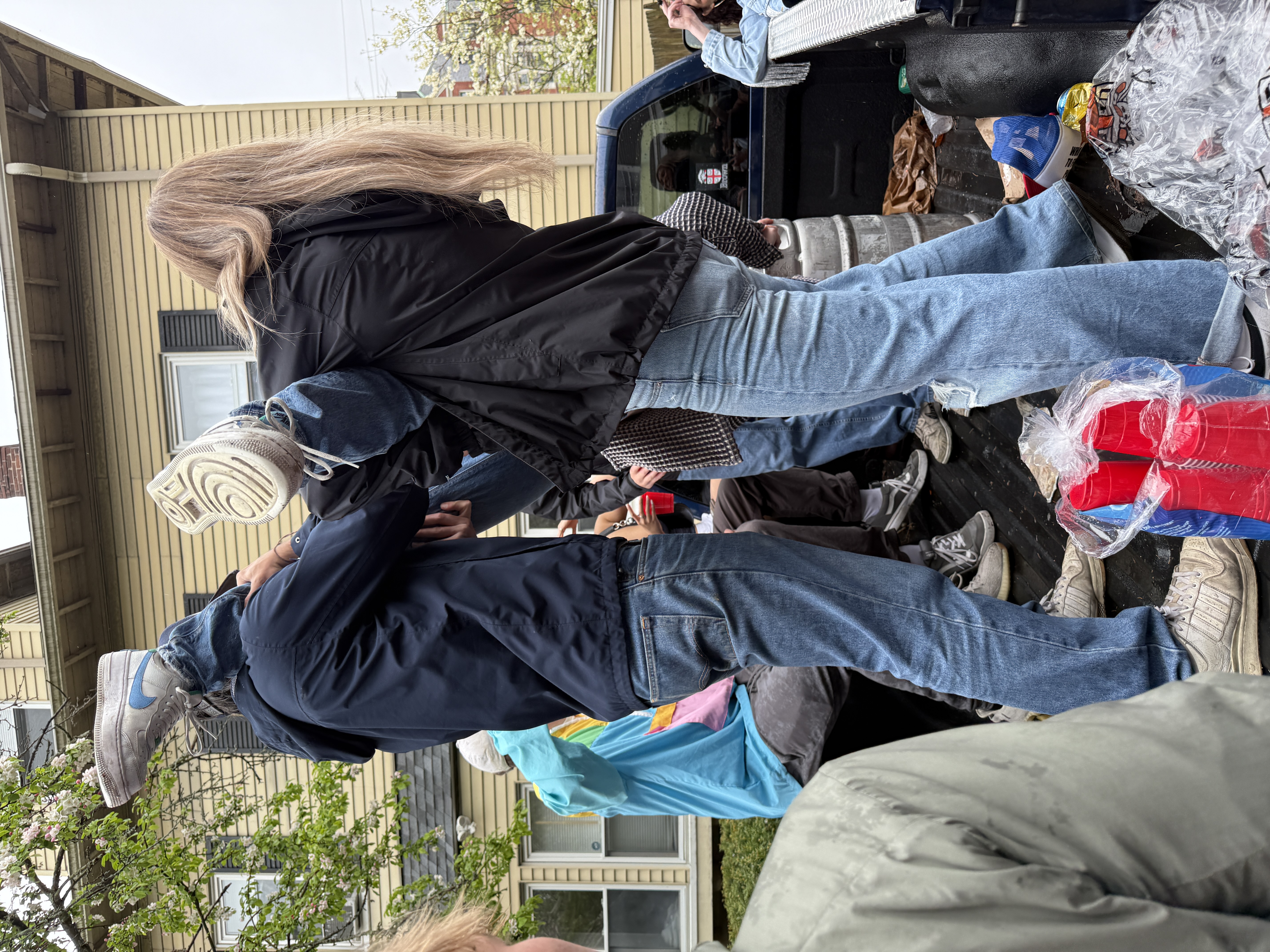 The image captures a lively outdoor scene where a group of young people seems to be engaging in playful antics, likely at a social gathering. In the foreground, one individual is lifting another, whose legs are playfully elevated, creating a sense of fun and camaraderie. The person being lifted appears to be enjoying the moment, with a joyful demeanor.

The background features a residential setting, characterized by a yellow house with distinct windows, suggesting a casual neighborhood atmosphere. The presence of friends in the background, some holding red cups, hints at a festive occasion, possibly a party or celebration. 

The overall vibe is lighthearted, reminiscent of college gatherings or casual get-togethers, where laughter and playful interactions are the norm. The scene evokes a sense of youthful exuberance, with the colorful cups adding a pop of vibrancy to the moment.