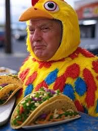 The image features a man in a vibrant chicken suit, complete with a large yellow beak and colorful feathers. His demeanor is serious, contrasting humorously with the absurdity of his costume, which is typically used for lighthearted promotions. The setting, likely a taco stand or food truck, suggests he’s involved in a quirky marketing campaign for tacos, adding to the comedic tone. Surrounding him are plates of tacos, brimming with fresh ingredients, emphasizing the food promotion aspect of the scene. This visual irony—an individual in a silly chicken outfit promoting delicious tacos—captures the essence of internet humor, reminiscent of popular memes that celebrate absurdity and playful marketing tactics.