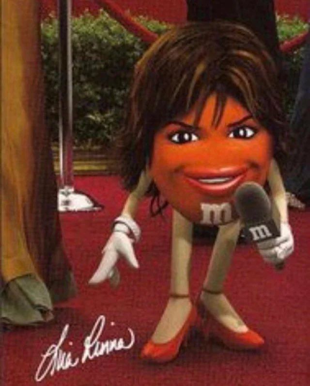 The image depicts an orange M&M character with human-like features, including a woman's hairstyle, makeup, and facial expression. The character is holding a microphone with the M&M logo on it and is standing on a red carpet, suggesting a celebrity or interview setting. The M&M is wearing high heels and white gloves. There is a signature in the bottom left corner that reads "Lisa Rinna," indicating that the character is meant to resemble or represent Lisa Rinna, a well-known actress and television personality. The image has been digitally altered to combine the features of Lisa Rinna with the anthropomorphic M&M character.