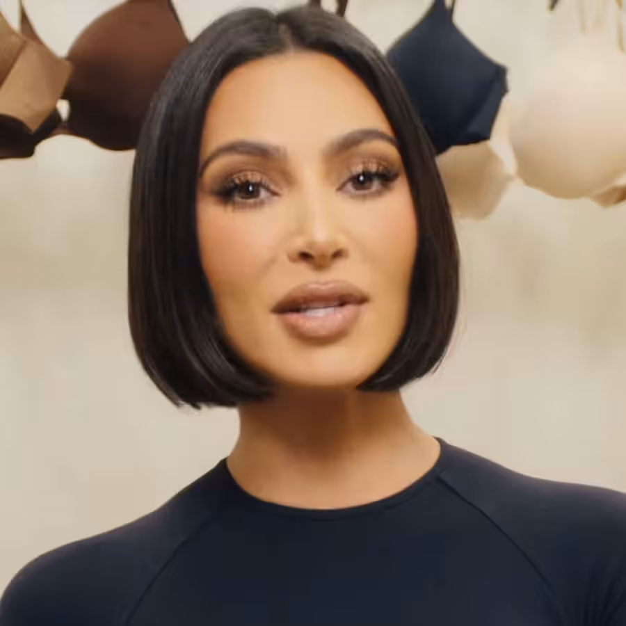 Kim Kardashian is captured in a close-up, looking directly at the viewer with a poised and professional demeanor, her lips slightly parted as if speaking. She sports a sleek, dark bob haircut and full, glamorous makeup, including defined brows and a nude lip. She wears a simple black long-sleeved top. The background, a light, neutral tone, features several bras in various shades of brown, black, and nude conspicuously hanging. This overt display of undergarments strongly indicates promotional material for her brand, SKIMS, with the bras serving as a direct visual cue to the product line. The humorous element lies in the almost comically direct 'bra-line' backdrop, leaving no doubt about the product being advertised.