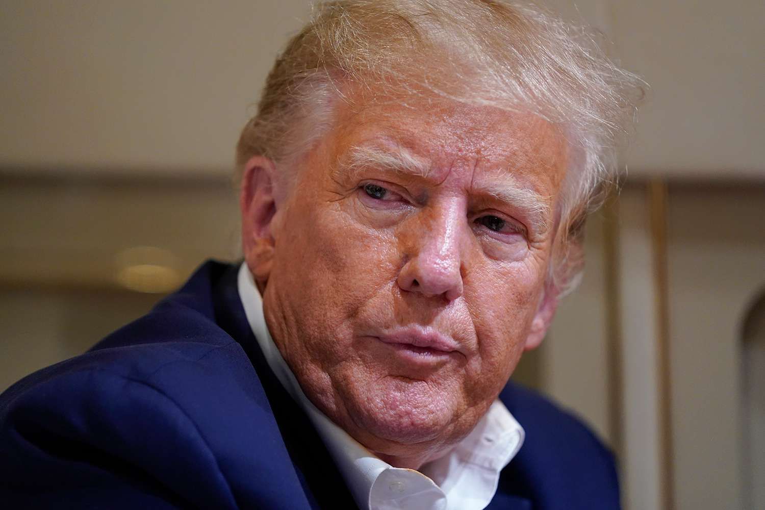 The subject, Donald Trump, is captured in a close-up, displaying a complex emotional state. His posture, though largely obscured, suggests a slight slump, with the blue suit jacket appearing somewhat rumpled at the shoulder. The white shirt, unbuttoned at the collar, adds a touch of informality, contrasting with the suit's formality and hinting at a moment of relaxation or dishevelment. His facial expression is characterized by slightly narrowed eyes, gazing upwards and to the left, conveying a sense of weariness or deep contemplation rather than direct engagement. The lips are subtly pursed or downturned, suggesting seriousness, perhaps mild disapproval, or a lack of overt enthusiasm. The blurred, neutral-toned background implies a private, possibly confined indoor setting. He projects an image of gravitas through his attire, yet his micro-expressions and slightly relaxed clothing communicate an underlying fatigue, introspection, or guardedness, revealing a tension between public persona and private emotional state.