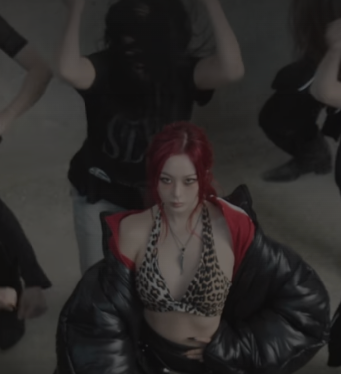 The image captures a woman with vibrant red hair and an intense, unyielding gaze, staring directly at the viewer. She sports a fierce leopard print halter top, a claw-like pendant, and a black puffer jacket draped over her shoulders, exuding a powerful, defiant demeanor. The blurry, dark figures in the background, some with arms raised, strongly suggest a dynamic performance or dance setting, characteristic of a K-pop music video. The dim, industrial backdrop enhances the edgy, dramatic vibe. Her focused expression amidst the chaotic energy behind her humorously implies she's the stylish, unbothered leader of a high-energy crew.
