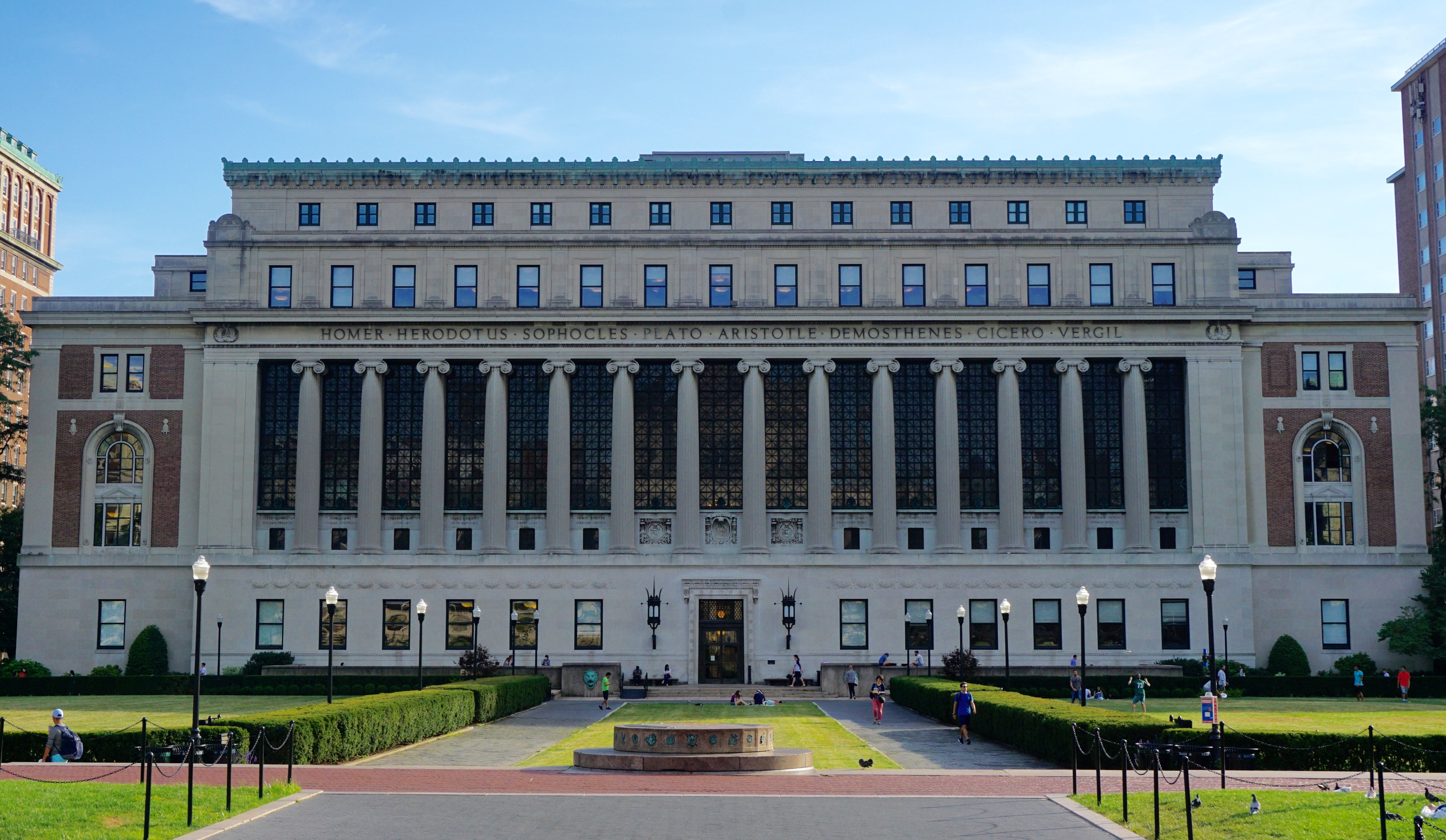 The image depicts the main library building of Columbia University, a distinguished Ivy League institution in New York City. The library's grand, neoclassical architecture features tall columns and an ornate facade, symbolizing knowledge and prestige. In the foreground, a well-manicured lawn is dotted with students, some lounging, others engaged in discussion, representing a vibrant academic atmosphere. The background showcases a clear blue sky, enhancing the library's stately presence. The scene conveys a sense of scholarly pursuit and community, with the library serving as a hub of learning and collaboration. The notable figures—Homer, Plato, and others—inscribed above the entrance reinforce the institution's commitment to intellectual heritage, creating a rich cultural context. Overall, the image captures the essence of academic life at Columbia, blending tradition with the energy of its diverse student body.