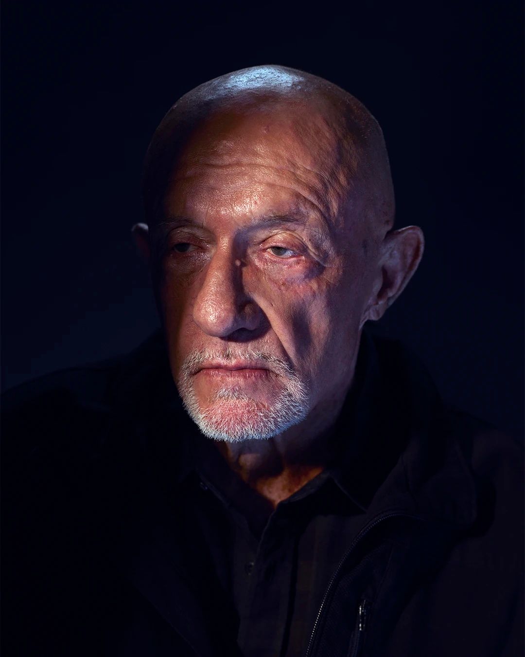 This striking portrait features Jonathan Banks as his iconic character, Mike Ehrmantraut, from 'Breaking Bad' and 'Better Call Saul'. His demeanor is intensely serious and world-weary, with a gaze that suggests deep thought or perhaps a silent judgment. The dramatic lighting, with a strong overhead light illuminating his bald head and grizzled beard against a stark, dark background, perfectly encapsulates the character's often grim and solitary existence. It's as if he's contemplating whether to take 'no half measures' or just wondering if he left the stove on. The minimalist background emphasizes his formidable presence, making him look ready to deliver a profound life lesson or simply ask for his payment.